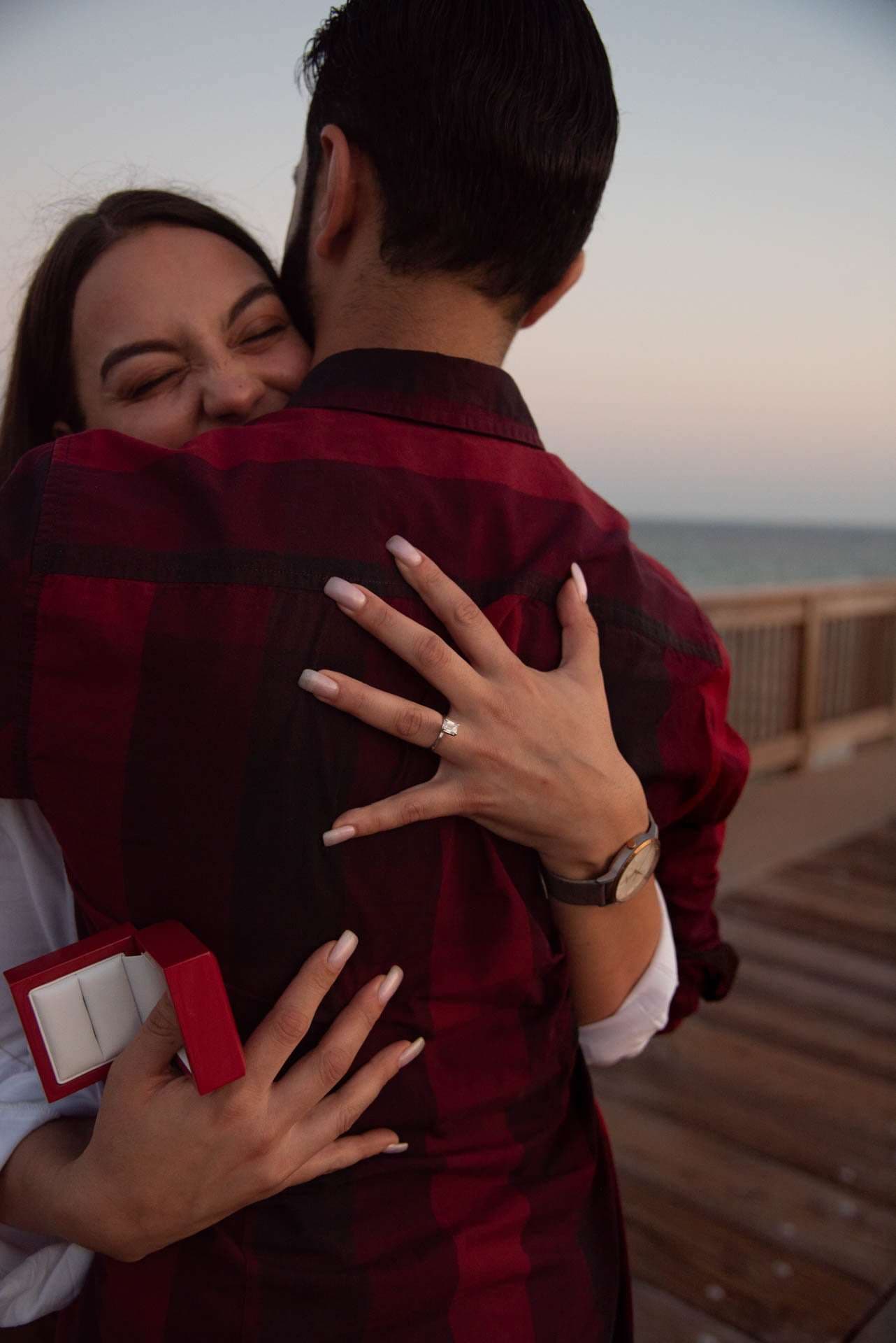 Woman smiling and hugging her fiancé, ring on her hand and open red ring box, on the Panama City Beach City Pier