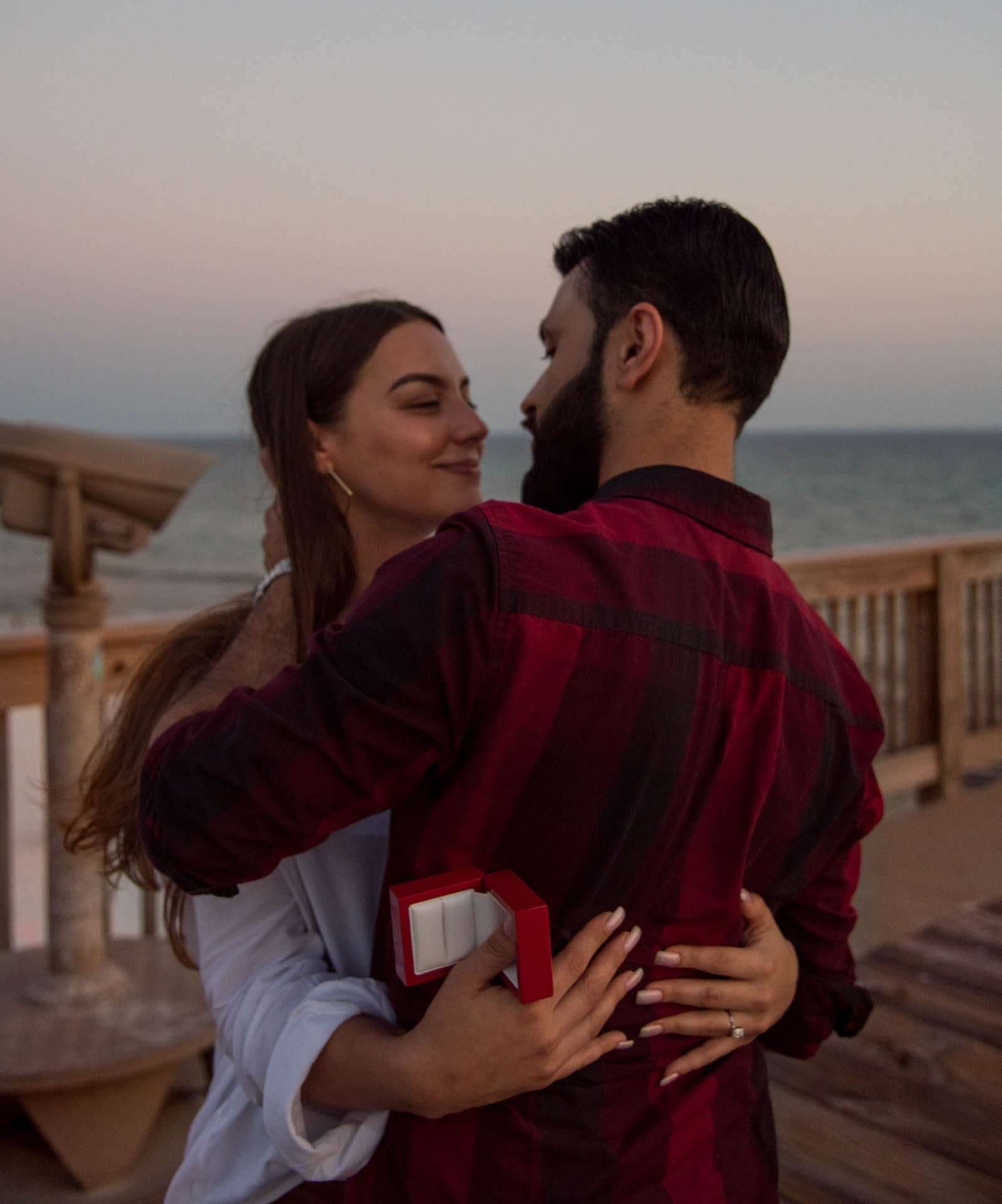 Woman hugging her new fiancé and holding an open red ring box on the pier at dusk