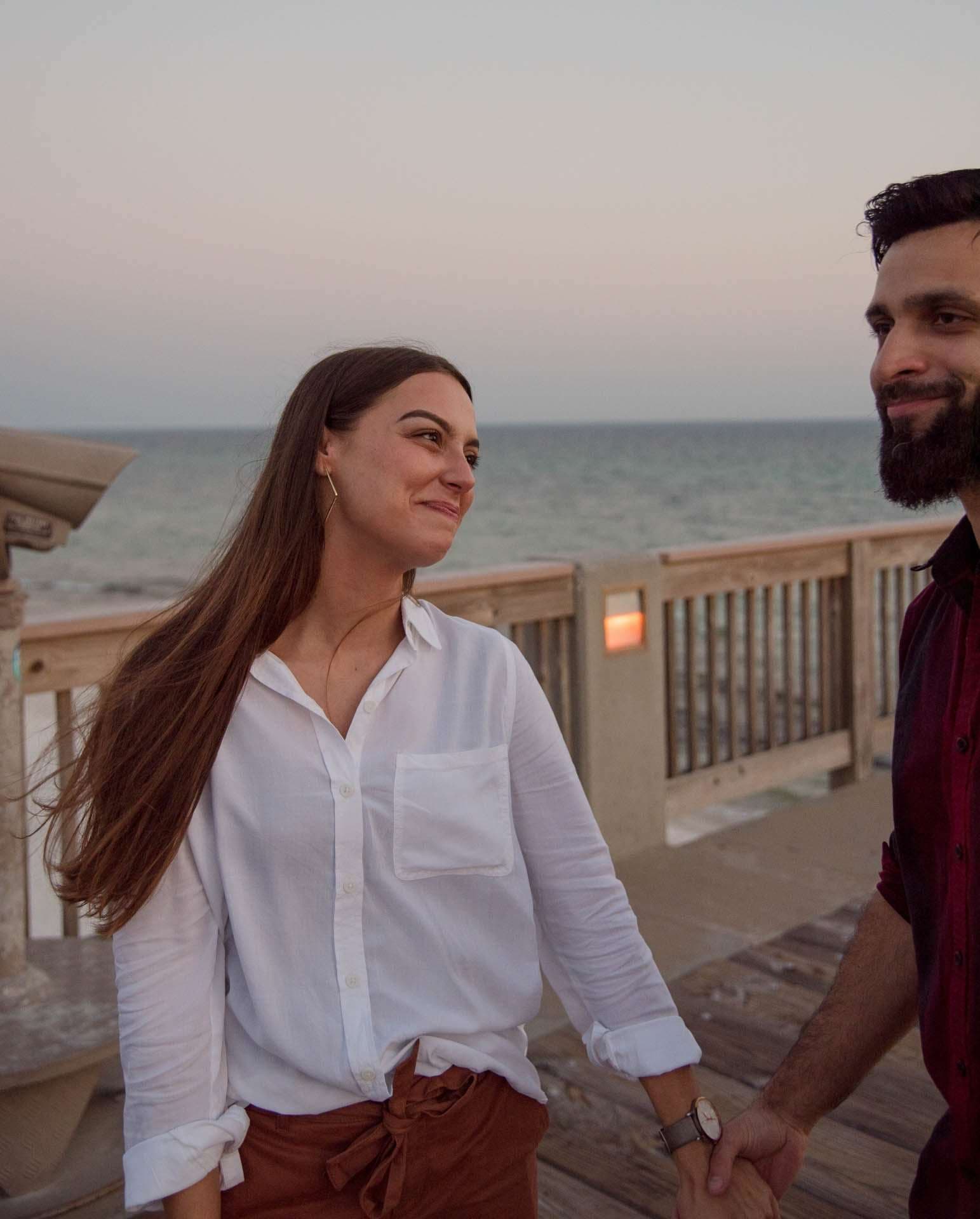 Smiling couple holding hands and walking along the Panama City Beach City Pier at dusk, gulf behind them