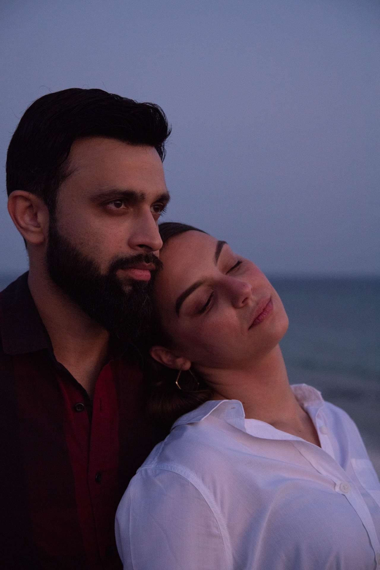 Bearded man in a dark shirt with a woman resting her head on his shoulder, eyes closed, by the sea at dusk