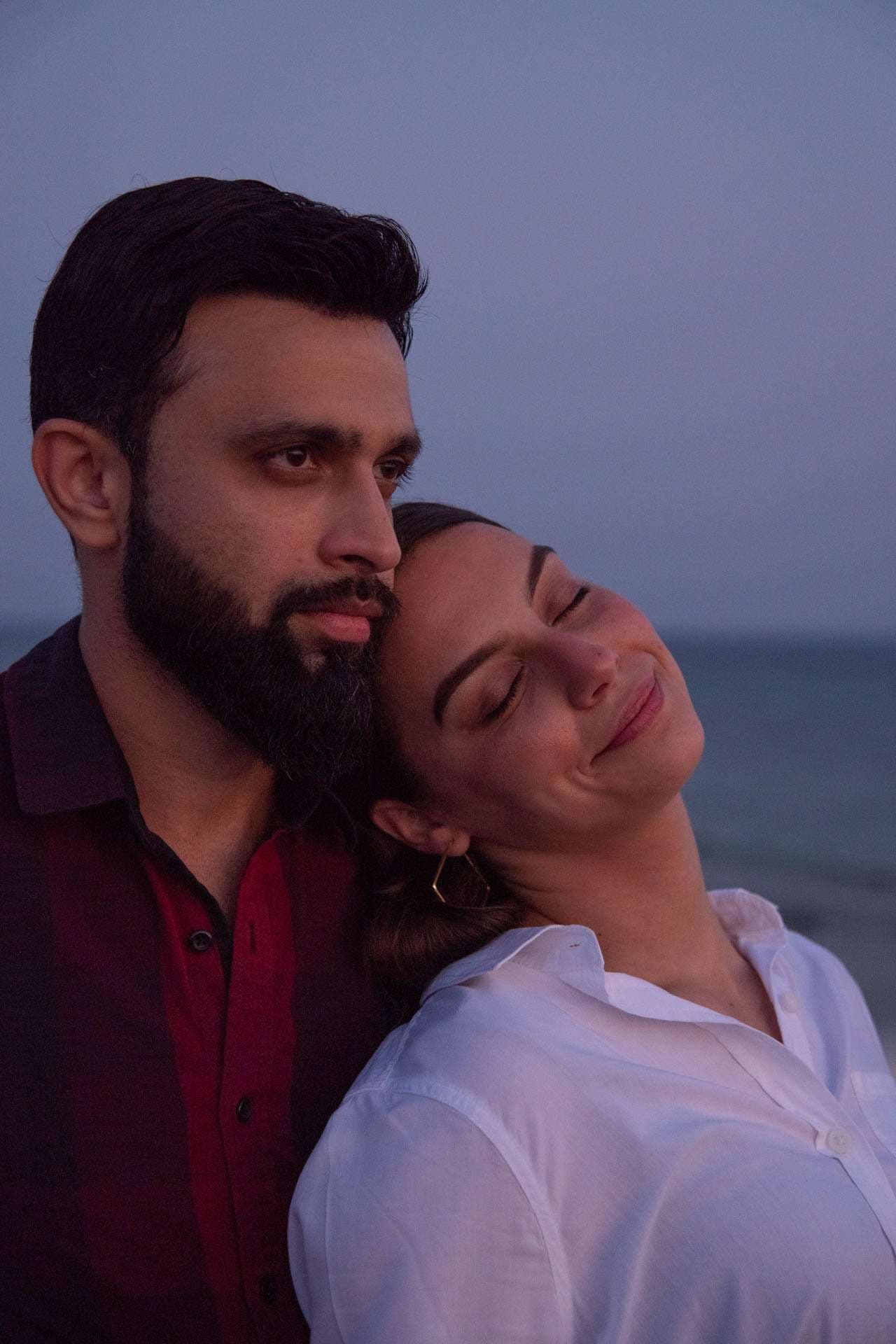 Bearded man in a dark red shirt with a woman resting her head on his shoulder, eyes closed, smiling at dusk by the water
