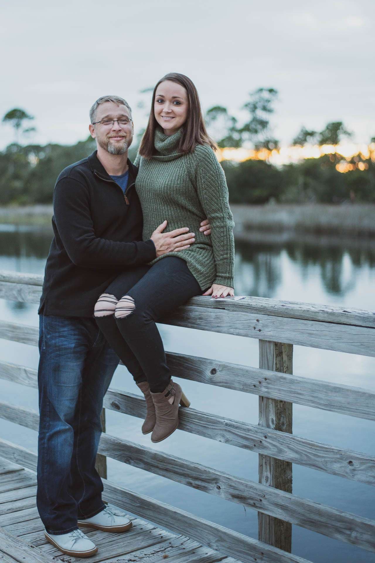 Smiling couple posing together on a wooden bridge railing at Camp Helen State Park, with a calm lake behind them