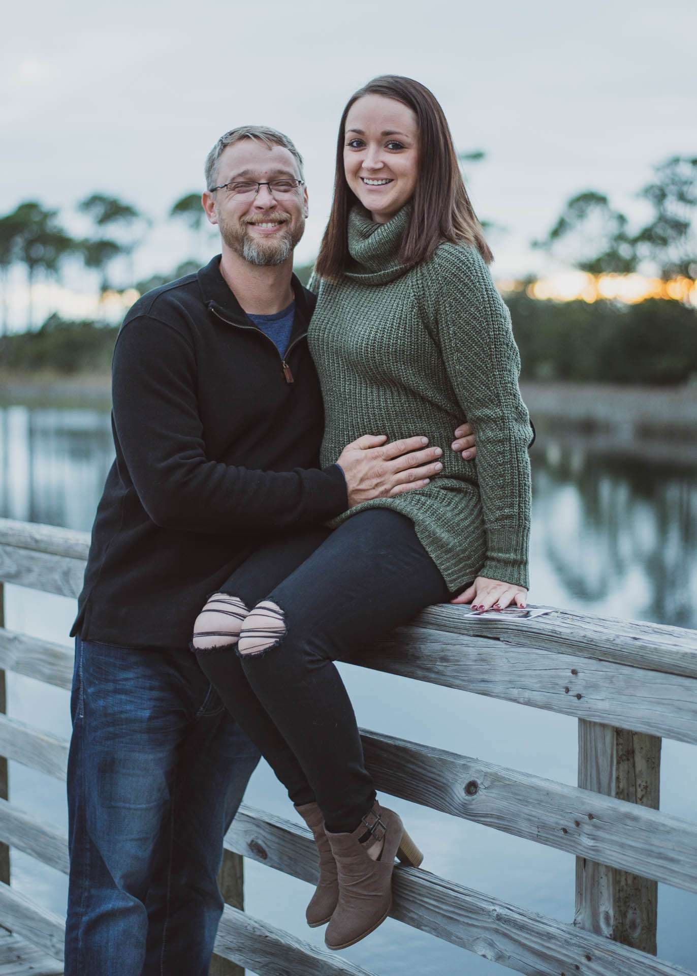 Smiling couple embracing on a wooden railing by a lake at dusk, his hand on her belly, at Camp Helen State Park