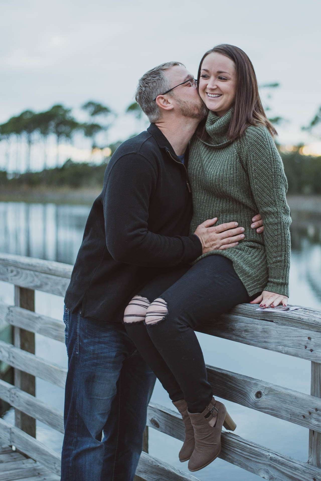 Man kissing smiling woman's cheek and resting his hand on her belly as she sits on a wooden railing by a lake
