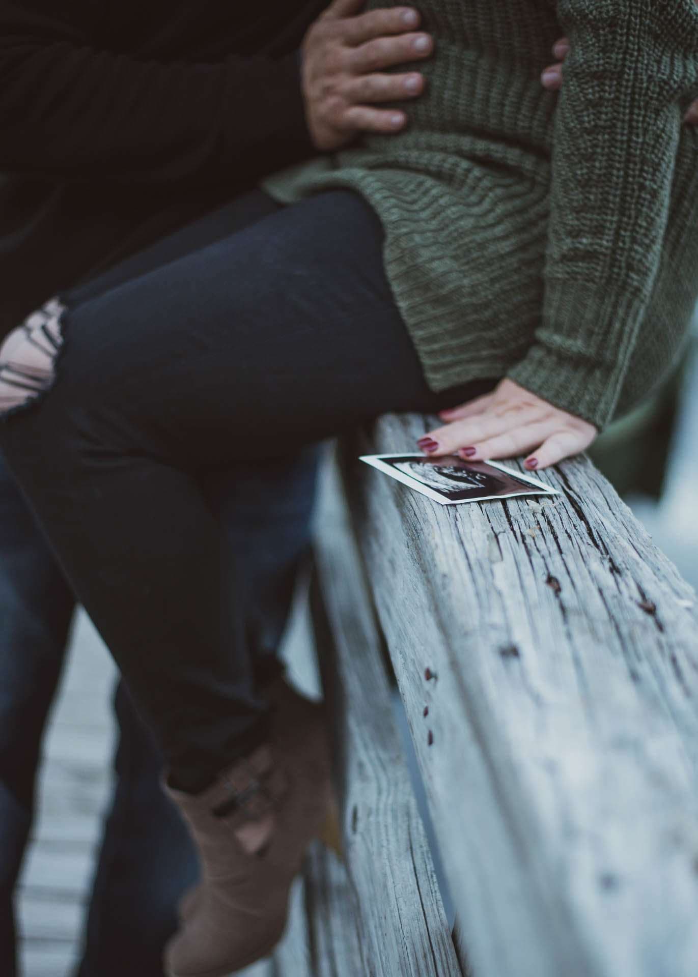 Couple embracing on a weathered wooden railing at Camp Helen State Park, an ultrasound photo resting beside her hand