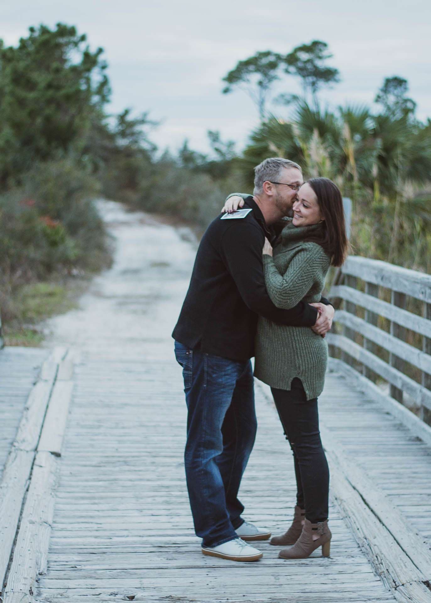 Couple embracing on a weathered wooden boardwalk at Camp Helen State Park, surrounded by coastal trees, woman smiling