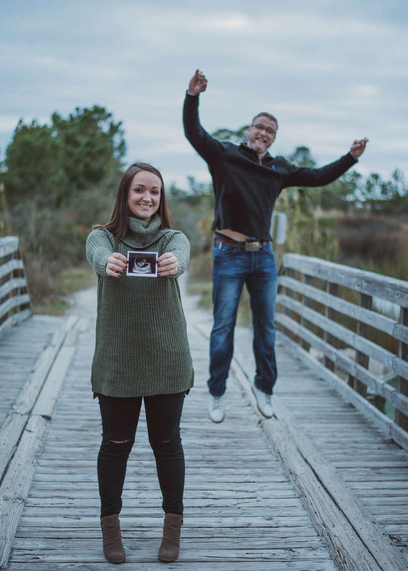 Smiling woman holding up an ultrasound photo on a wooden boardwalk while a man leaps joyfully behind her