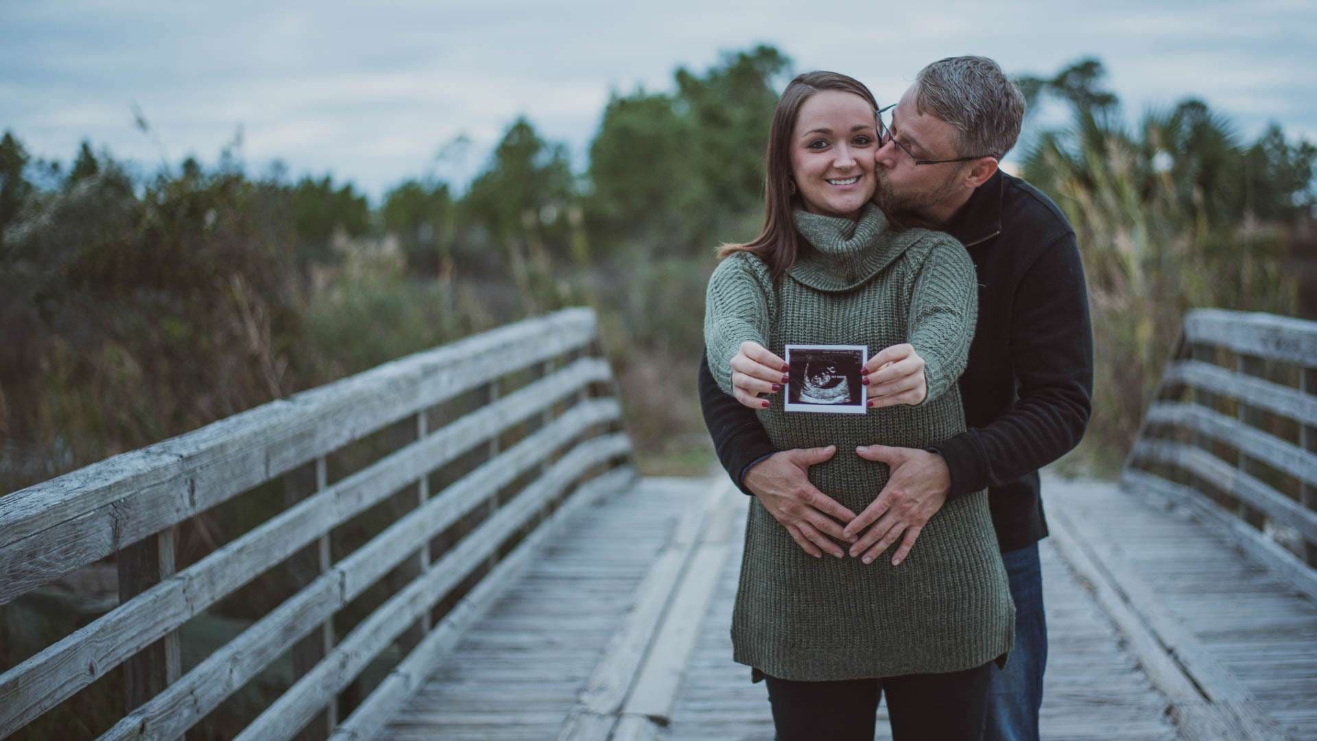 Man kissing his smiling partner's cheek as she holds an ultrasound picture over her baby bump on a wooden boardwalk