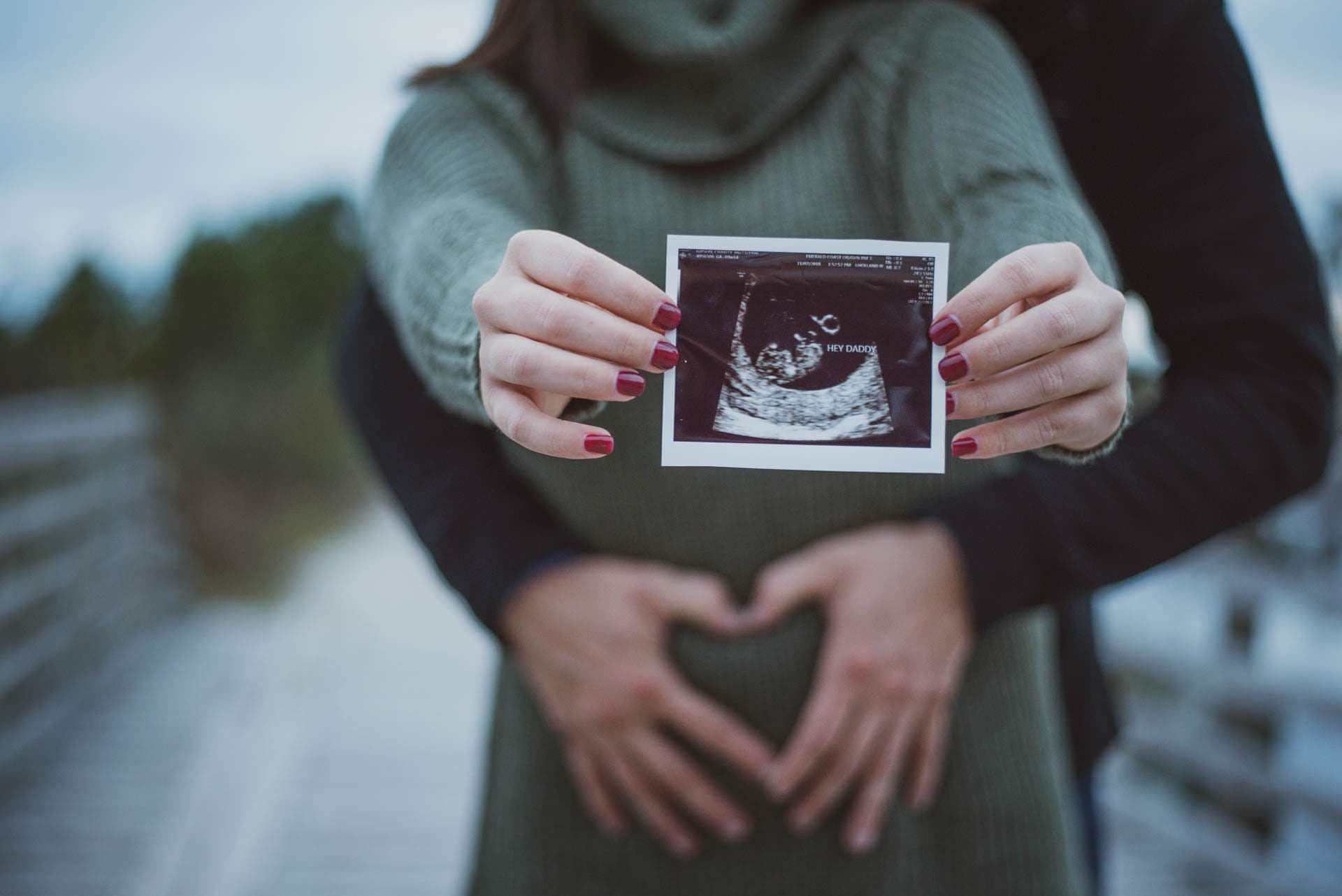 Expectant couple forming a heart over the baby bump while holding up an ultrasound photo on a wooden boardwalk