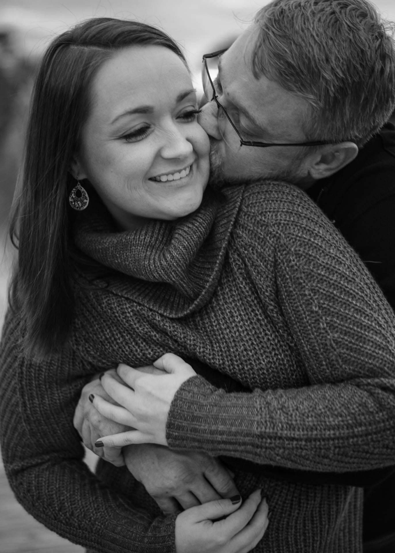Black-and-white portrait of a man in glasses kissing the cheek of a smiling woman in a chunky knit sweater