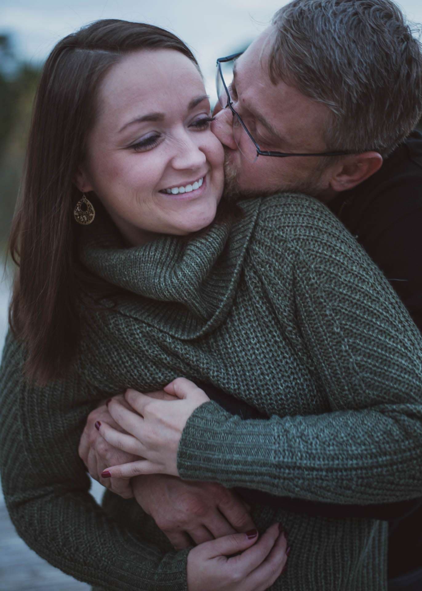 Smiling woman in a green knit sweater being hugged and kissed on the cheek by a bearded man in glasses