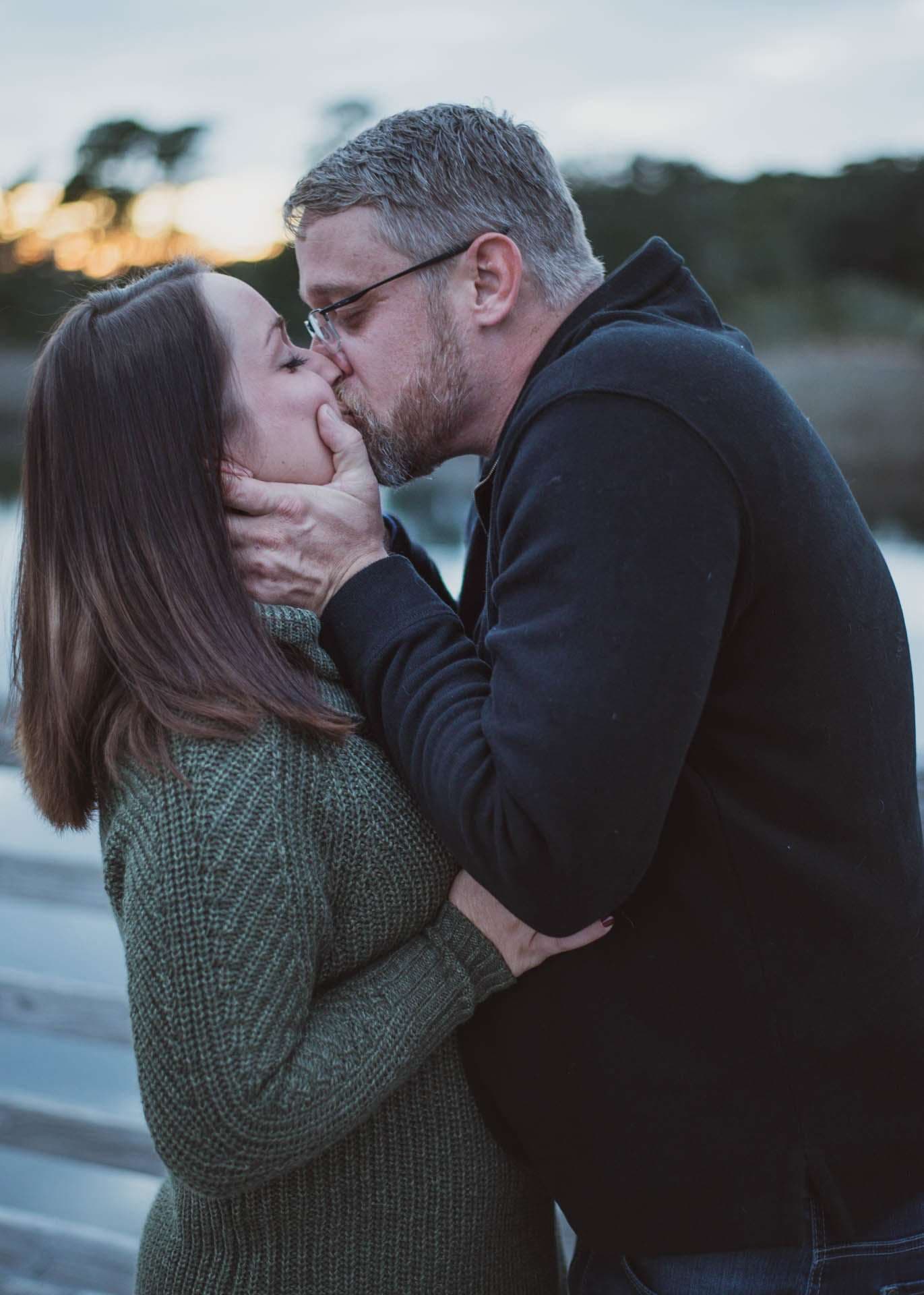 Couple kissing at dusk on a wooden boardwalk by the water, she in a green knit sweater, he in a dark jacket