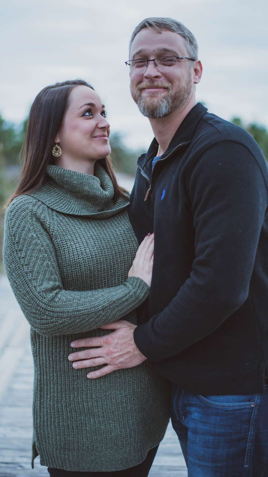 Smiling bearded man in glasses embraces a woman in a green knit sweater as she gazes up at him, hand on her belly
