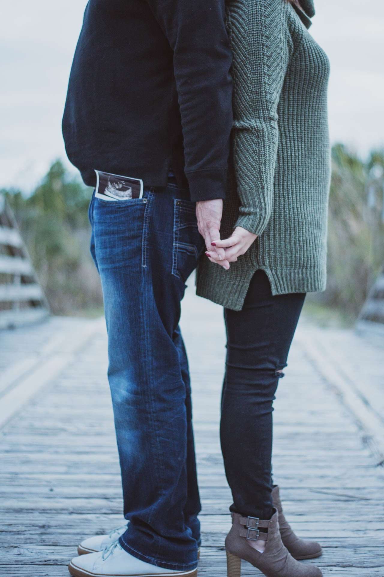 Couple holding hands on a wooden boardwalk at Camp Helen State Park, an ultrasound photo tucked in his back pocket