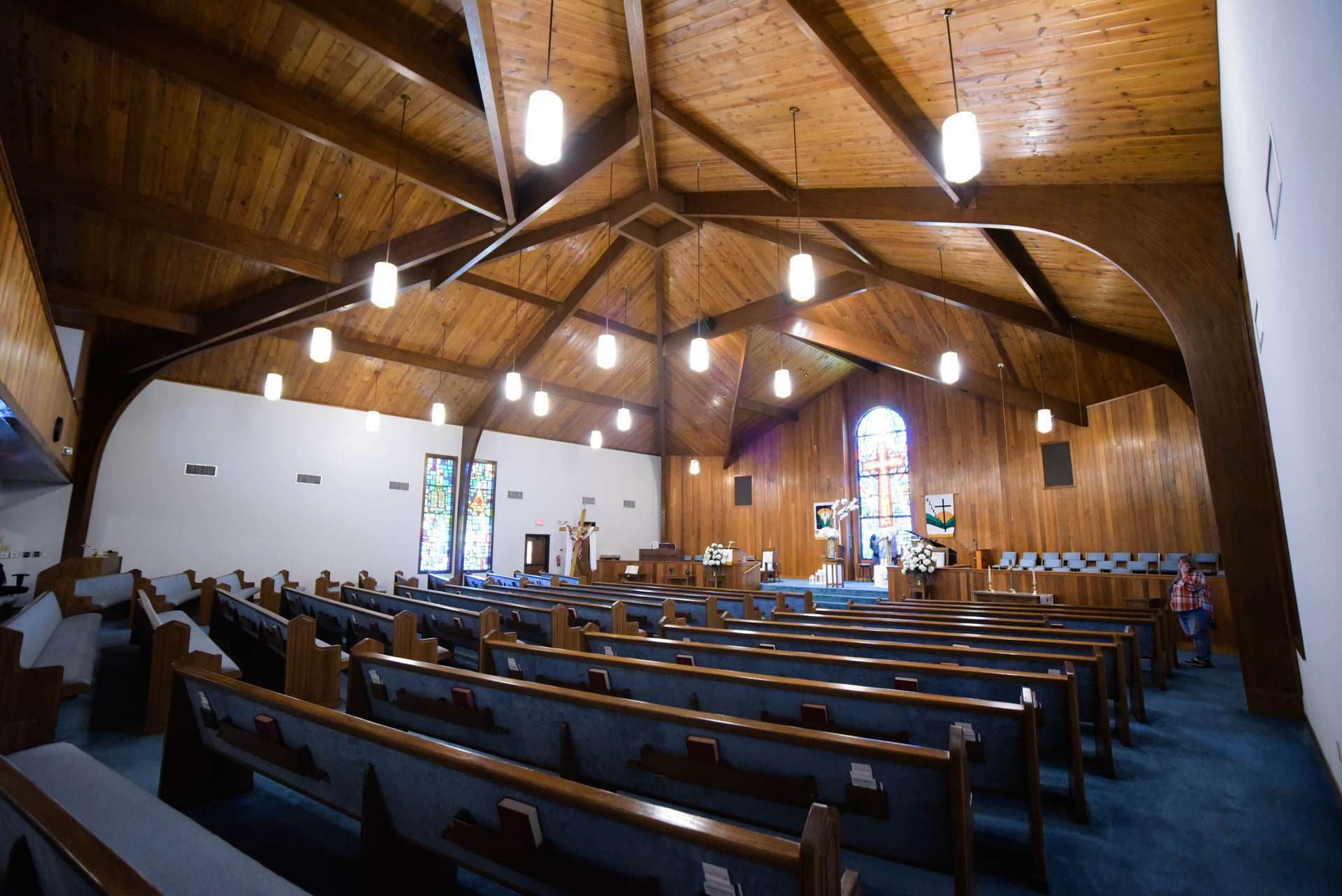 Empty church sanctuary with wooden pews, vaulted wood-beam ceiling, pendant lights and a stained glass window at the front