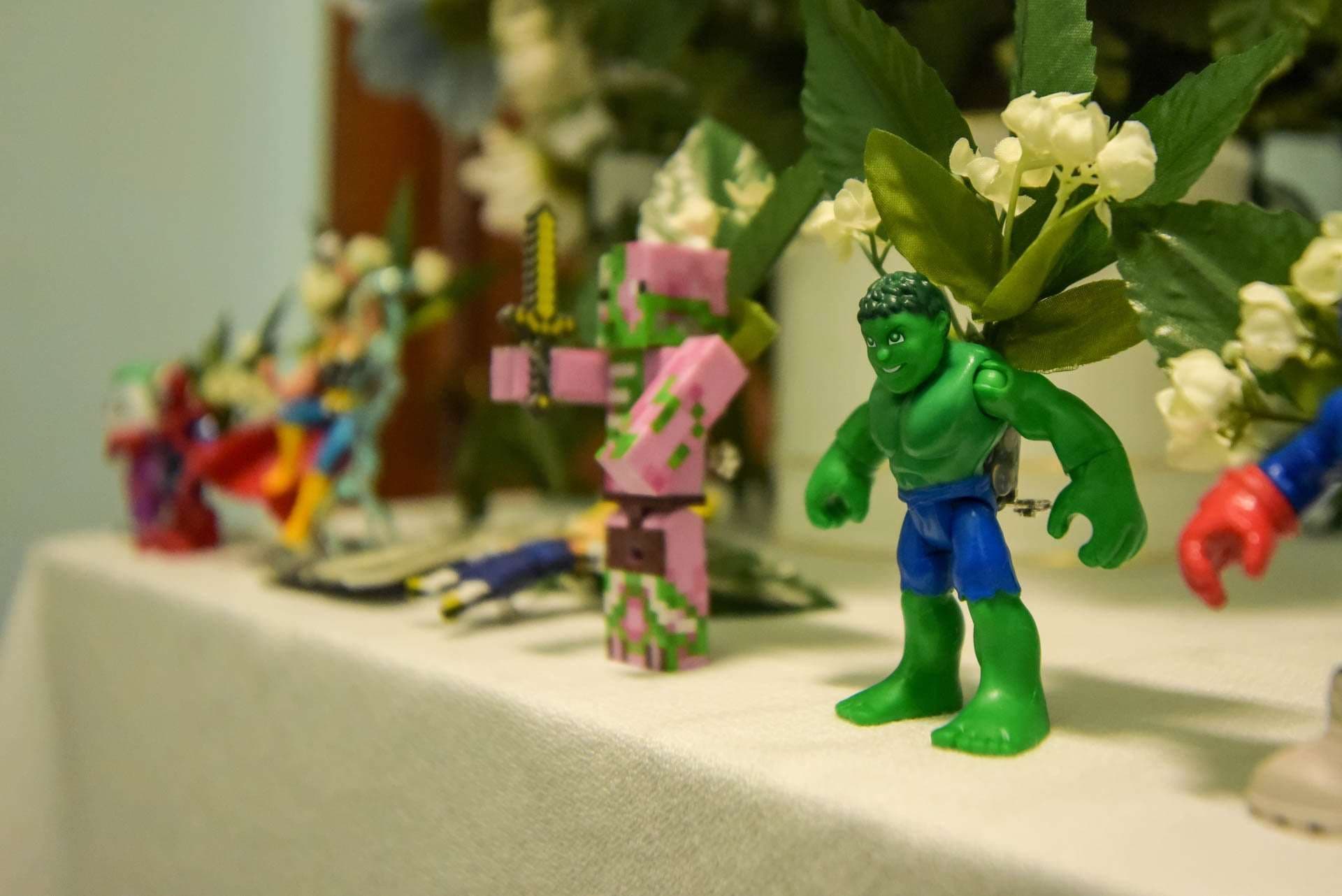 Hulk and Minecraft zombie piglin toys lined up with other superhero figures on a white tablecloth beside white flowers