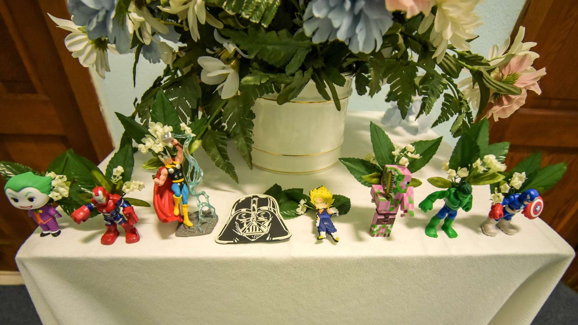 Superhero and Darth Vader figurines with small white flowers on a white-draped table beneath a floral arrangement