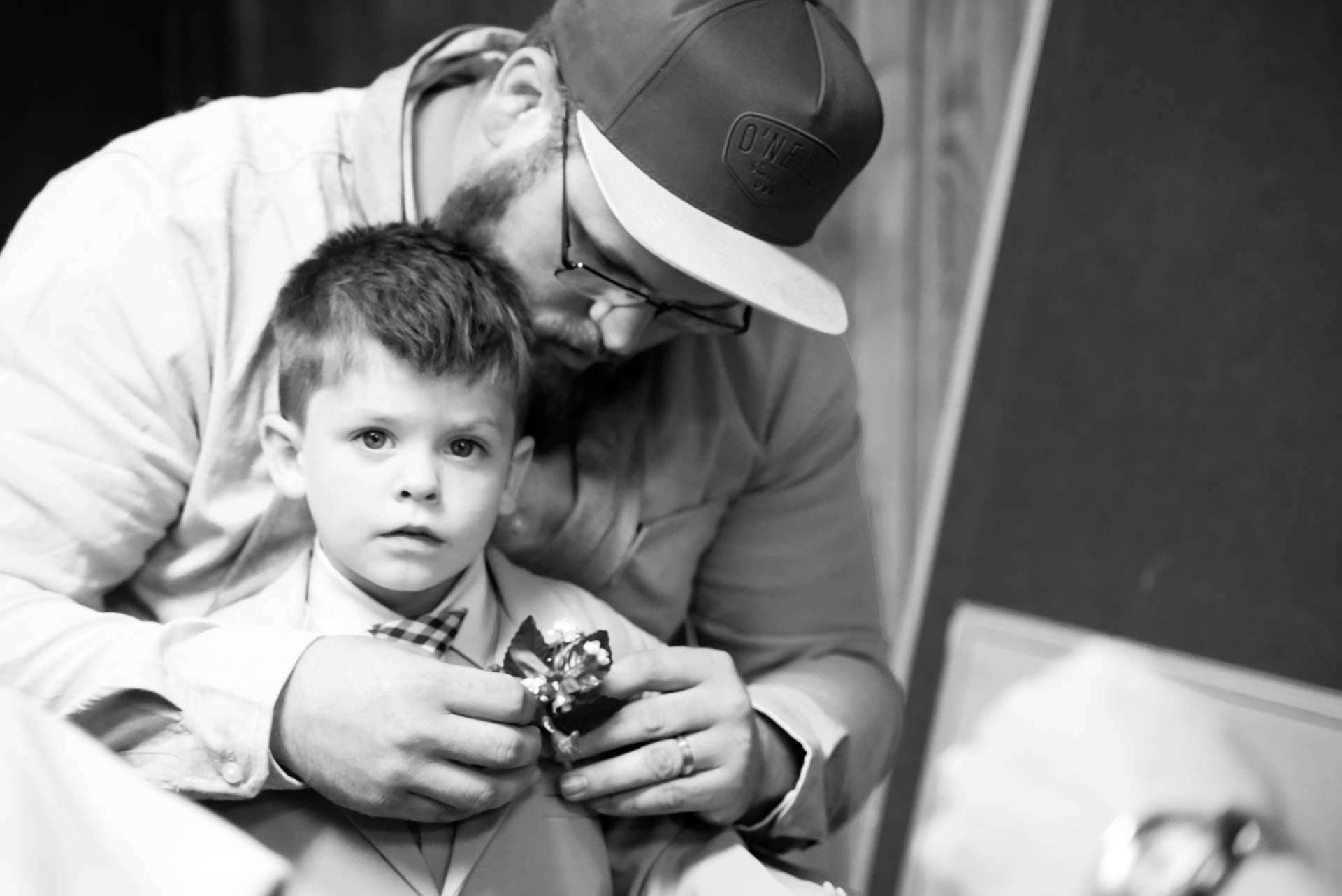 Black-and-white shot of a bearded man in a cap pinning a boutonniere on a young boy's suit jacket
