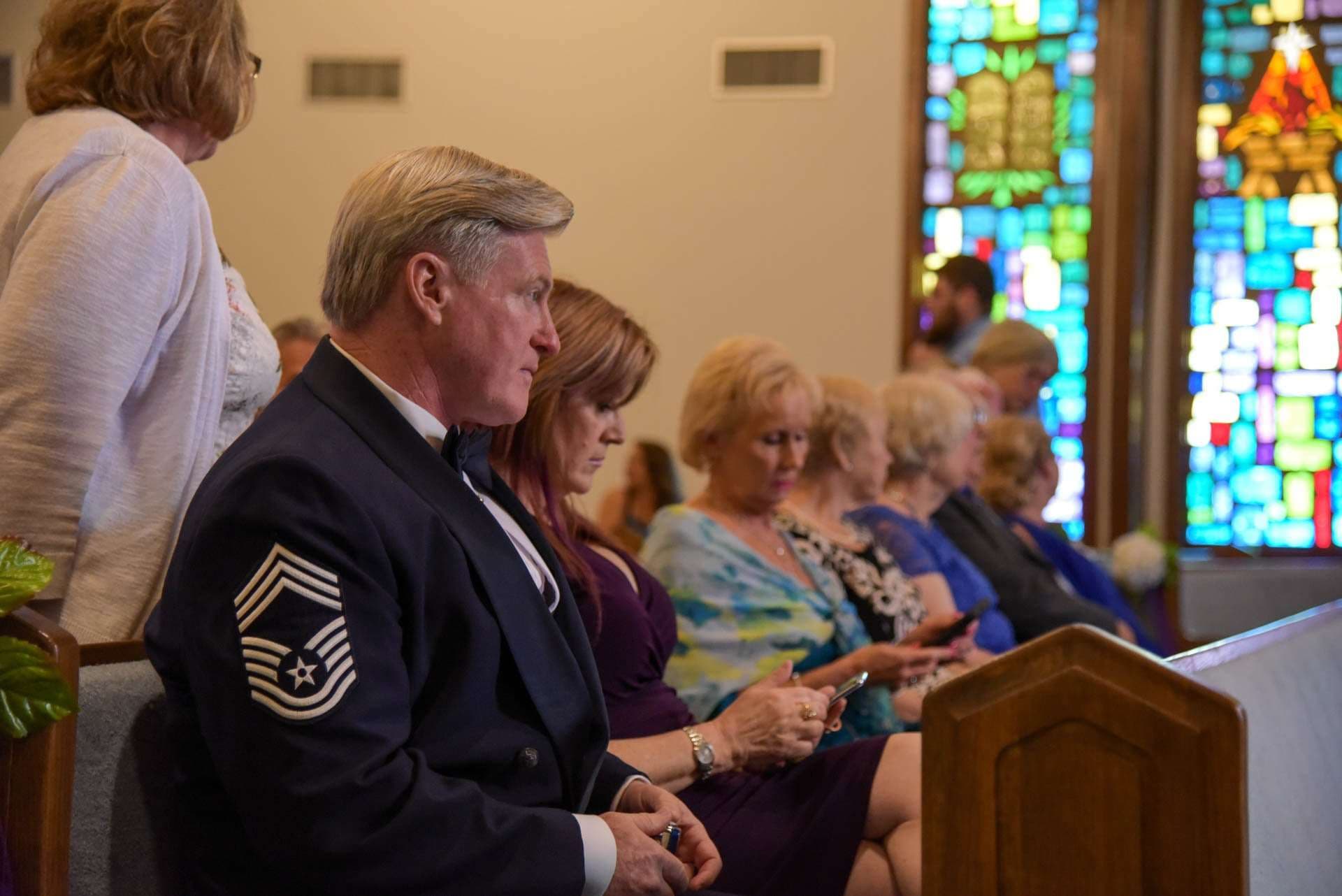 Man in dark military dress uniform sits in a church pew beside guests looking at phones, stained glass behind