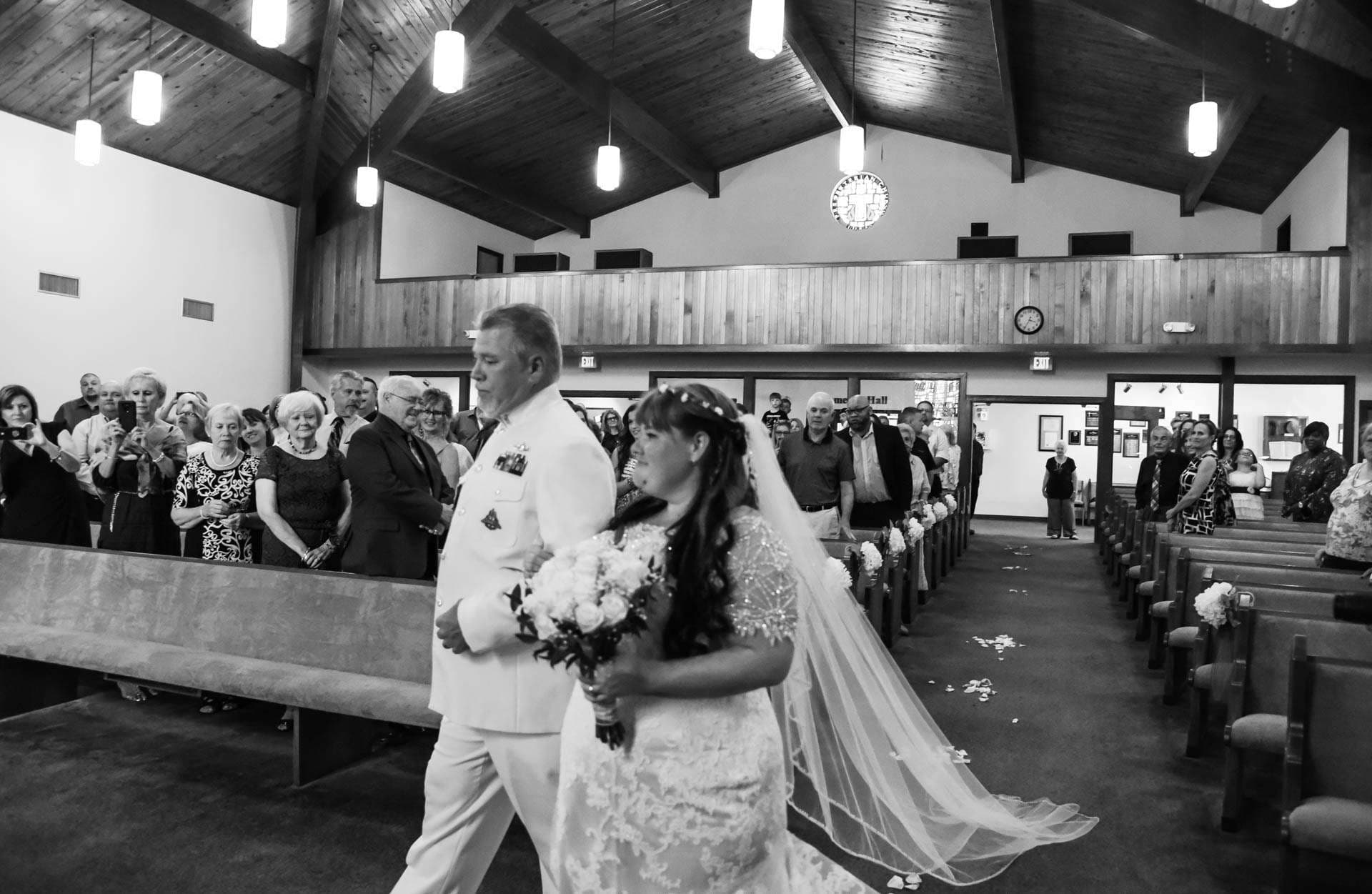 Black-and-white shot of a bride in a lace gown with veil walking down a church aisle beside a man in a white uniform