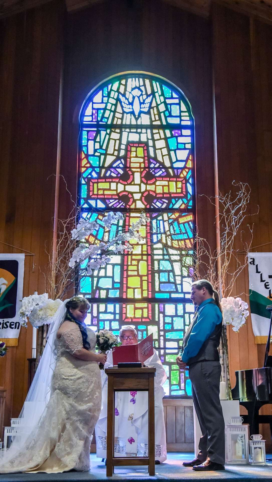 Bride in a lace gown and veil and groom in a teal shirt stand before an officiant in front of a stained glass cross