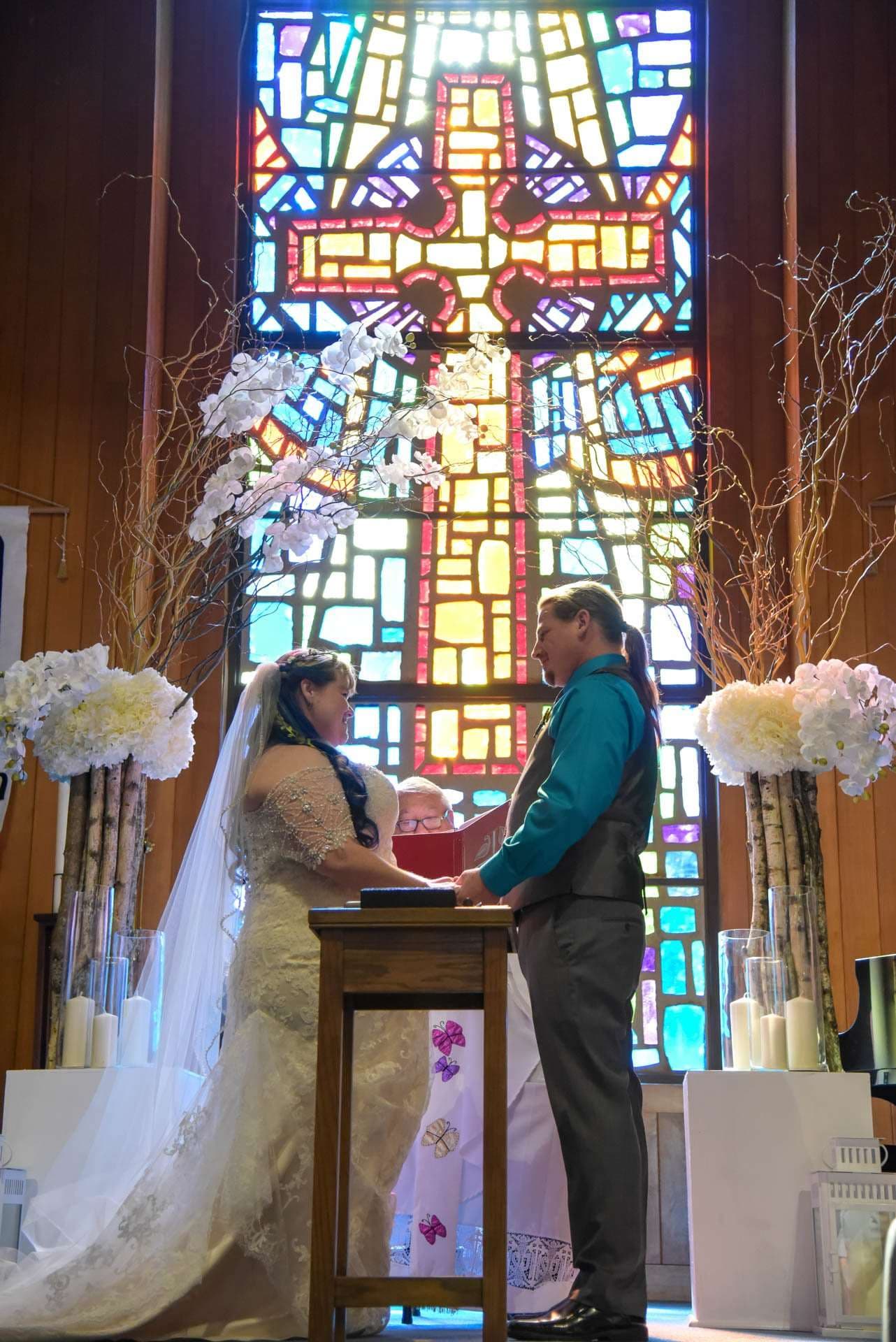 Couple exchanging vows at a wooden podium before a large stained glass window, flanked by white floral arrangements