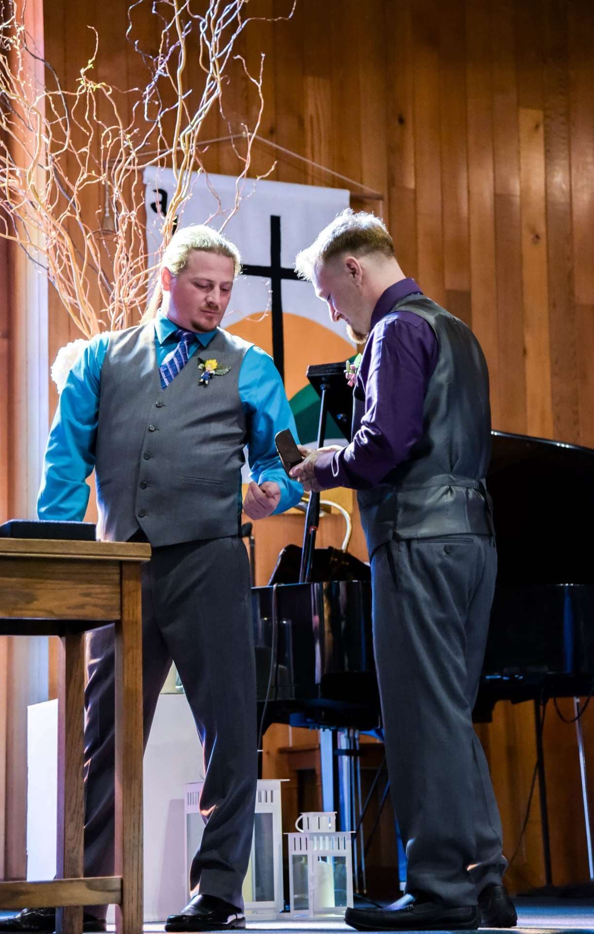 Two men in gray vests and dress shirts stand facing each other near a piano during a church wedding ceremony