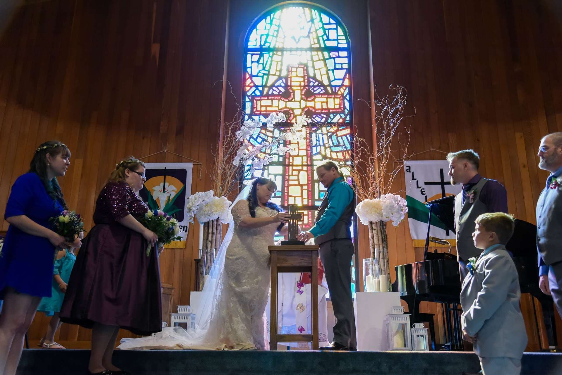 Couple at a small wooden table during a church ceremony beneath a stained glass cross, with attendants and a boy watching