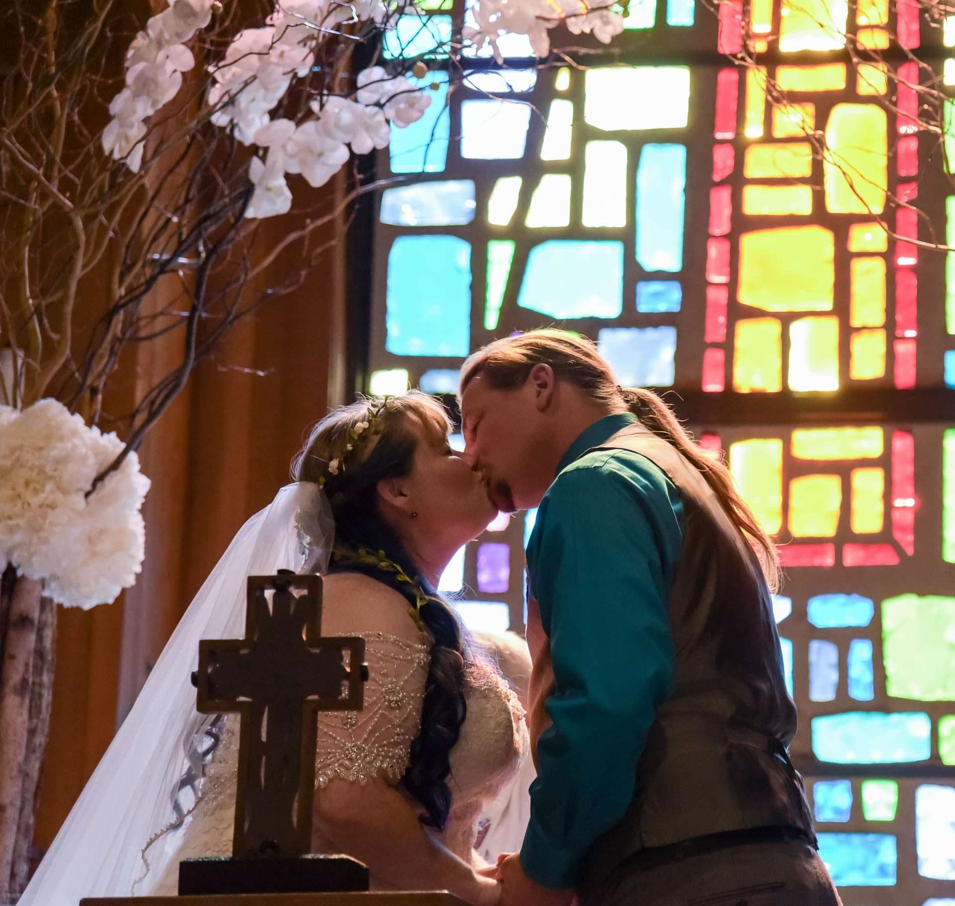 Couple kissing at a church altar beside a wooden cross, with colorful stained glass glowing behind them