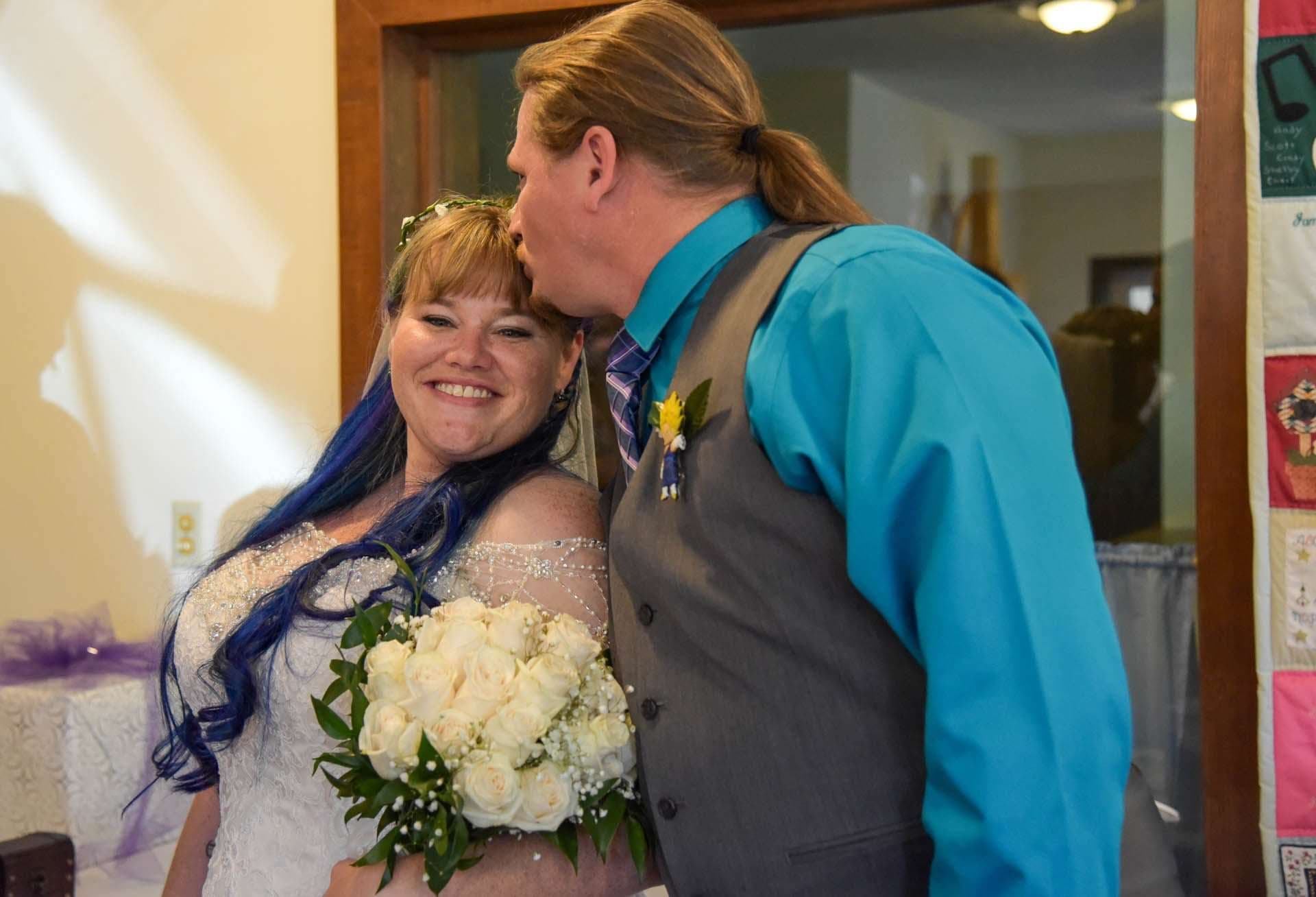 Smiling bride with blue-streaked hair holding a white rose bouquet as her partner in teal shirt kisses her forehead