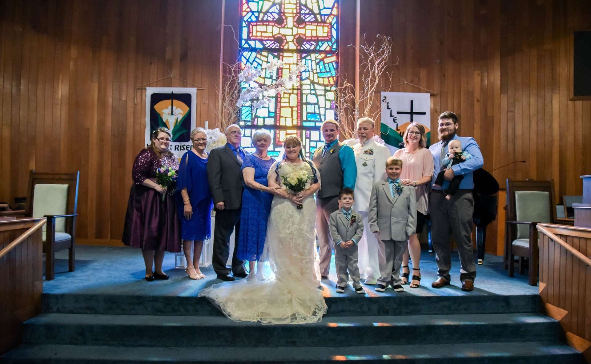Bride holding a bouquet poses with family, including two boys in gray suits, on church steps before a stained glass window