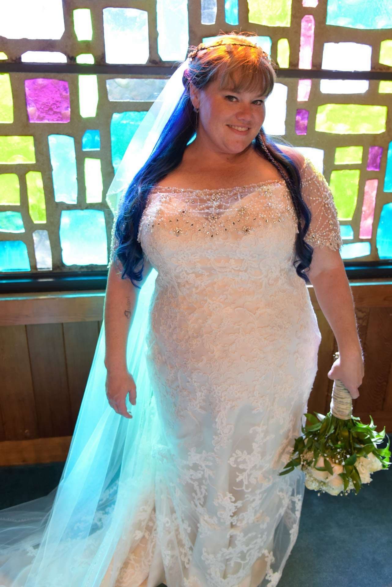 Smiling bride with long blue hair in a beaded lace gown and veil holding a bouquet before stained glass windows
