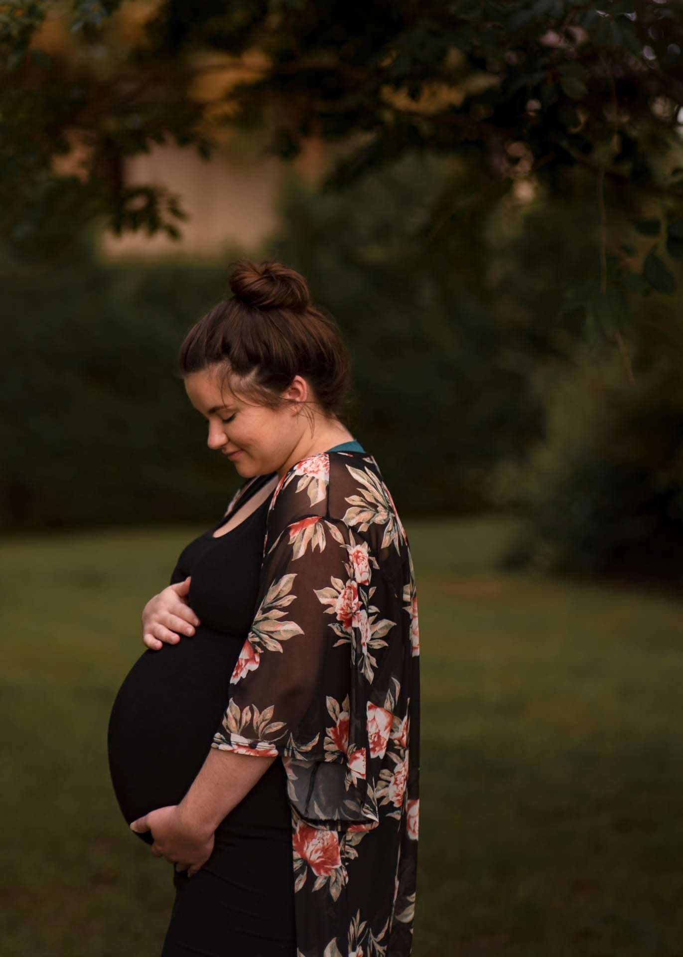 Pregnant woman in a floral kimono cradling her belly and smiling down, standing on a grassy lawn among trees