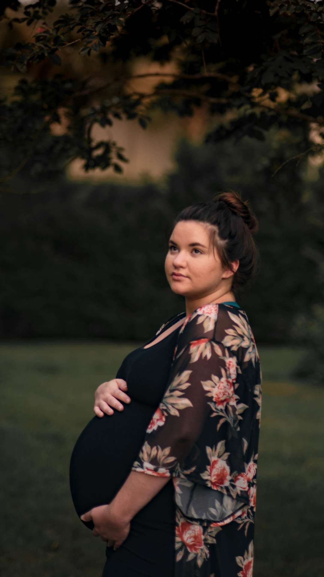Pregnant woman in a floral kimono cradling her belly and gazing away in a grassy, tree-shaded setting at dusk