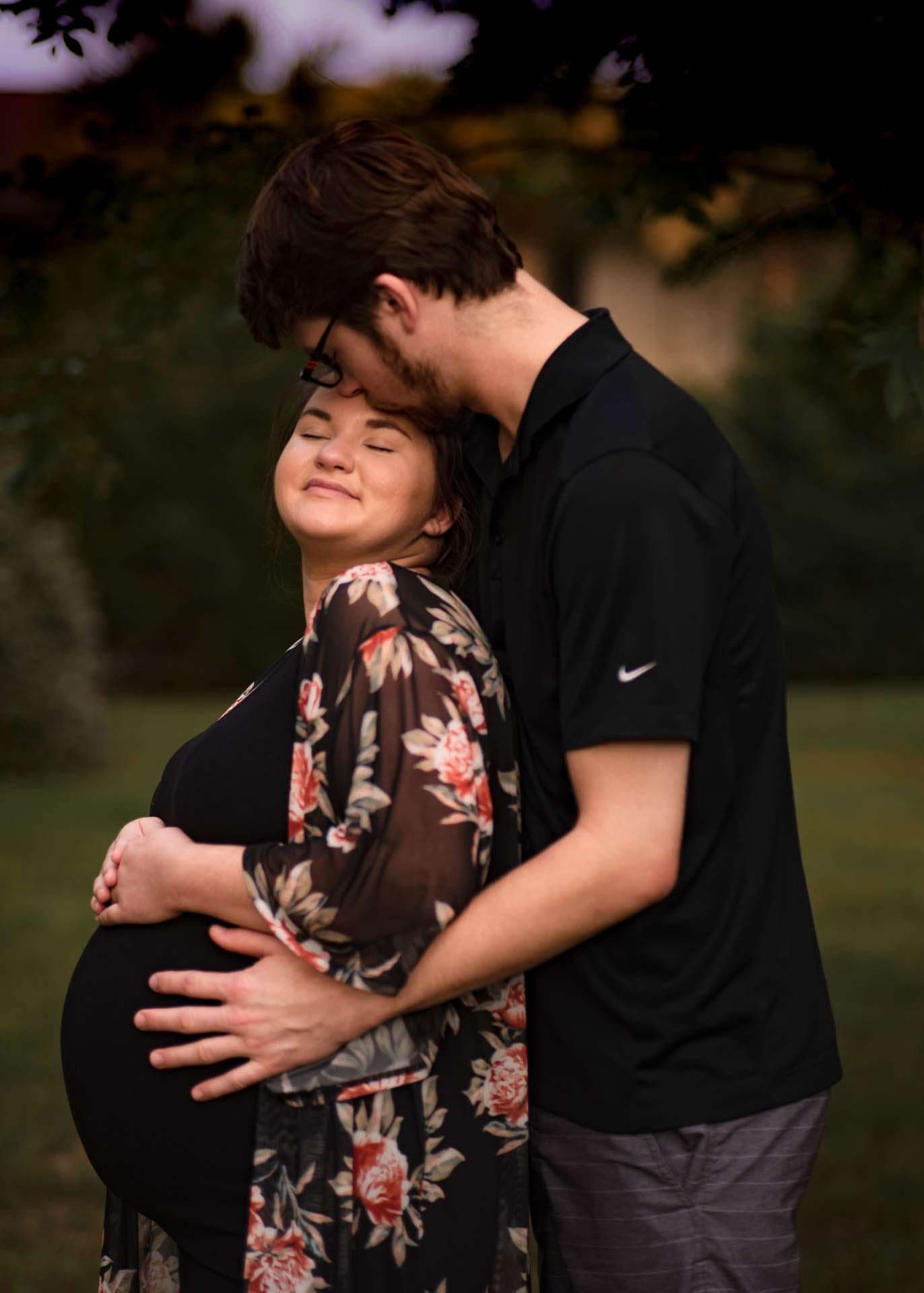 Man kissing the forehead of his smiling pregnant partner, hands on her belly, on a grassy lawn at dusk