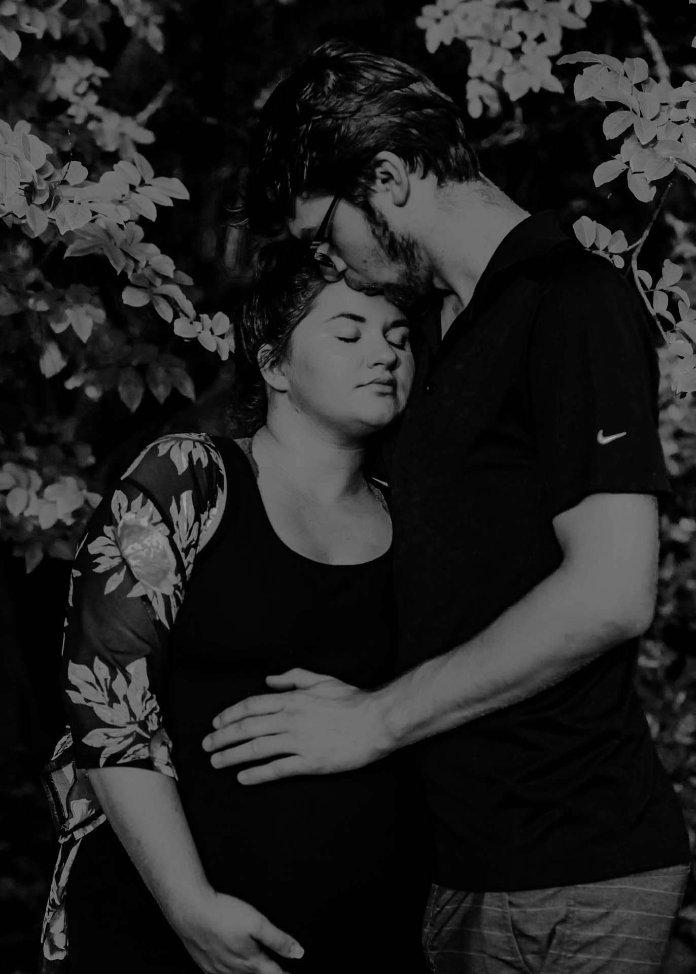 Black-and-white portrait of a man kissing his pregnant partner's forehead, hand on her belly, among leaves