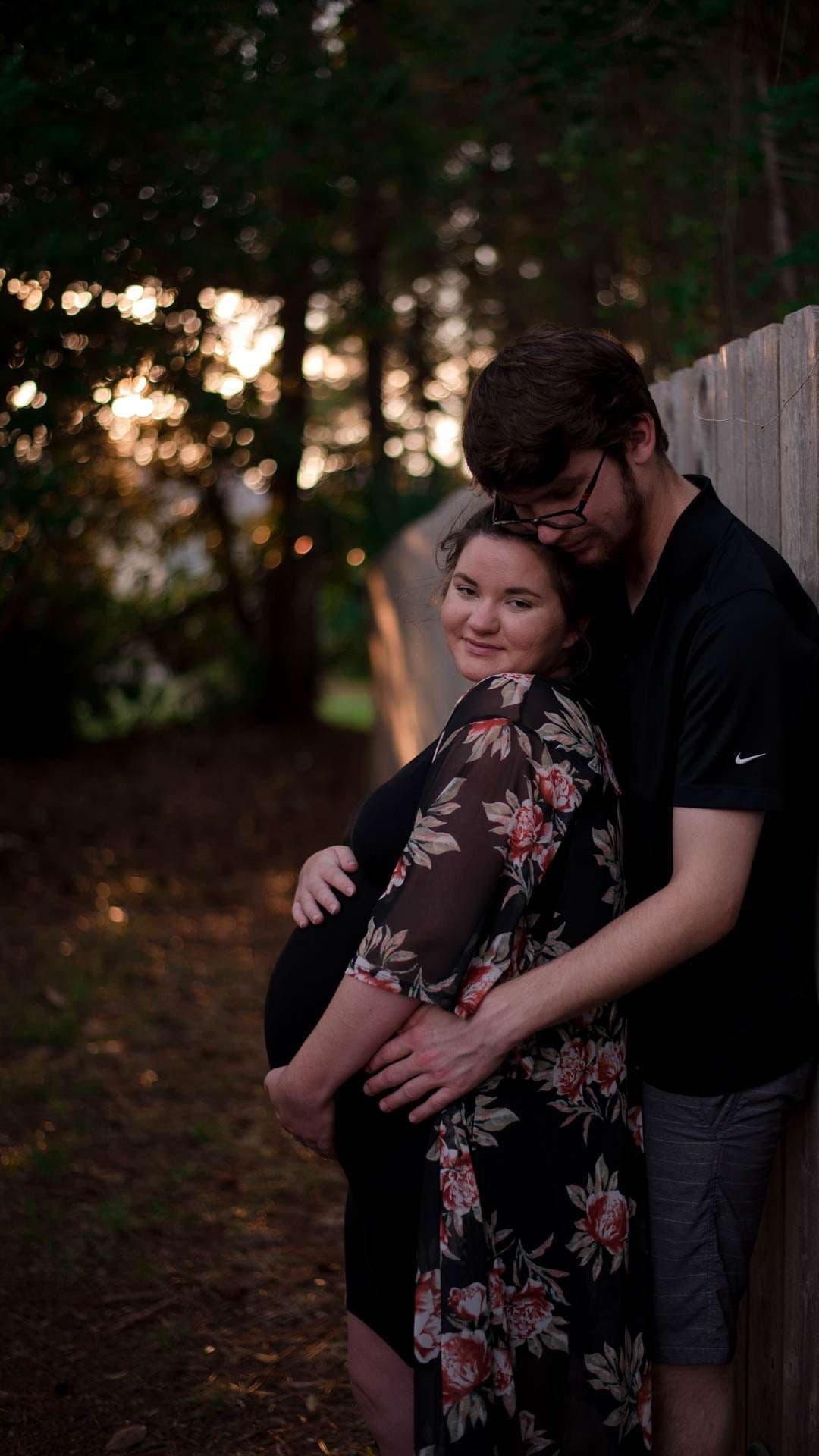 Expectant couple embracing beside a wooden fence in the woods, his arms wrapped around her baby bump at sunset