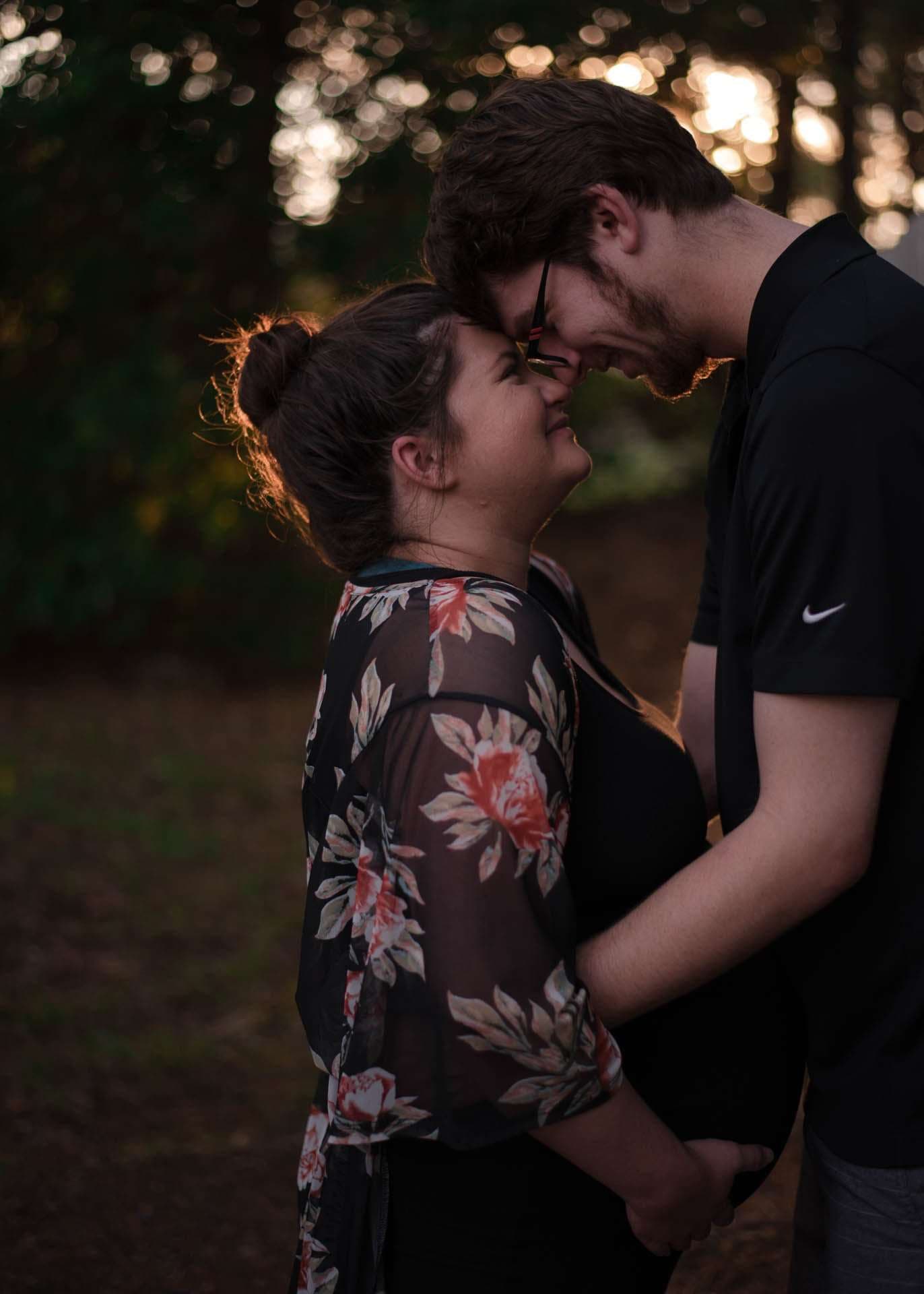 Couple touching foreheads and smiling in the woods at dusk, her hand resting on her baby bump, she wears a floral kimono
