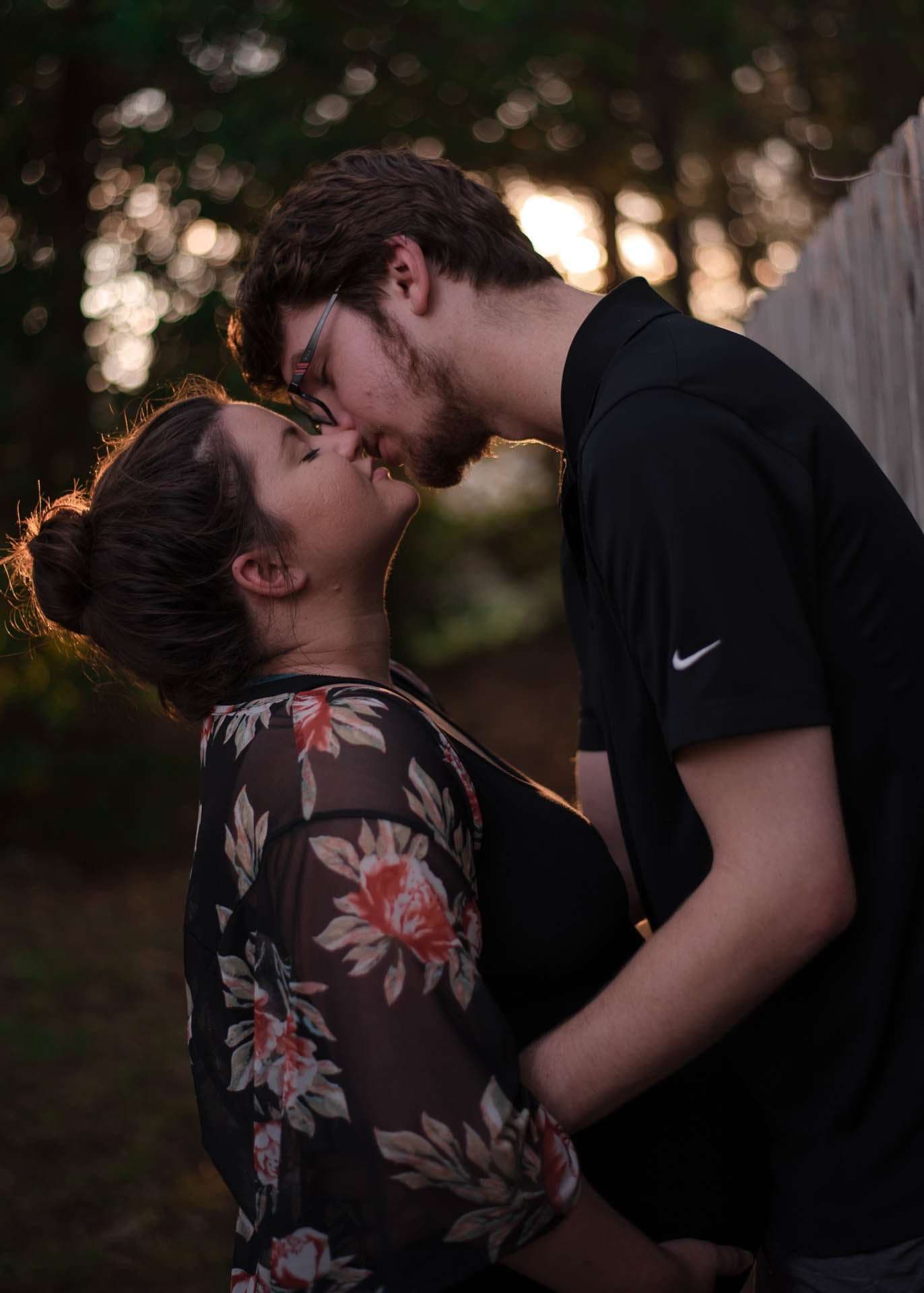 Couple about to kiss in a backlit wooded setting at sunset, she in a floral sheer top, he in a black polo