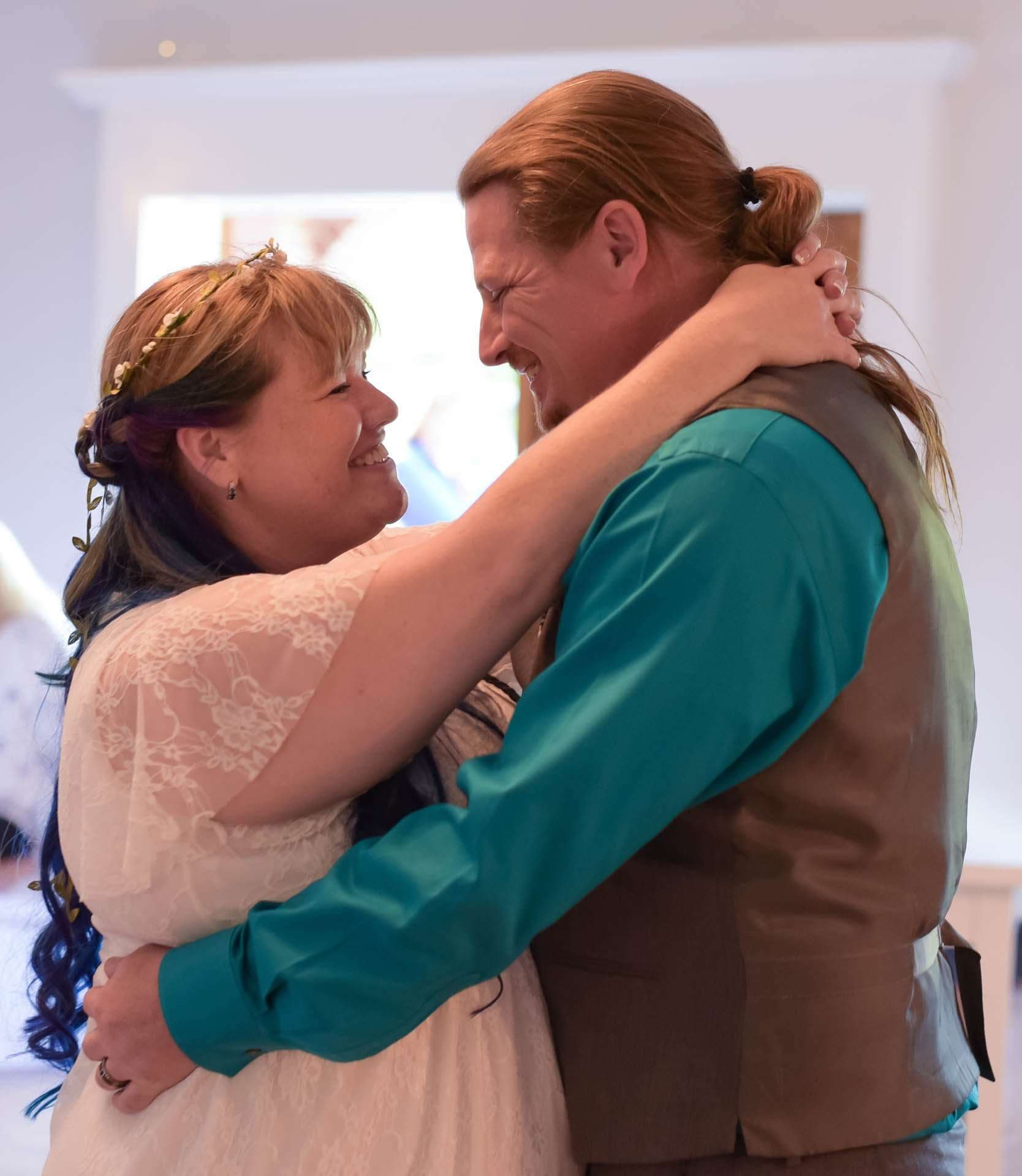 Smiling couple embracing during a slow dance, she in a white lace dress with a flower crown, he in a teal shirt and vest