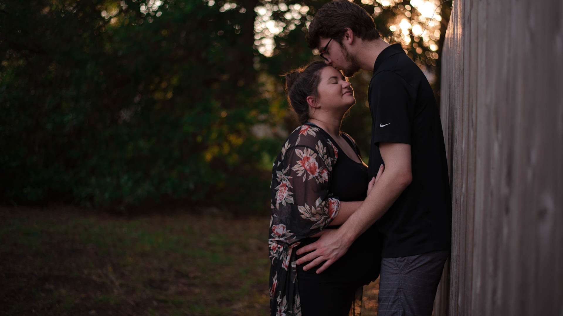 Man kissing his partner's forehead as she smiles with closed eyes, leaning against a wooden fence near trees at sunset