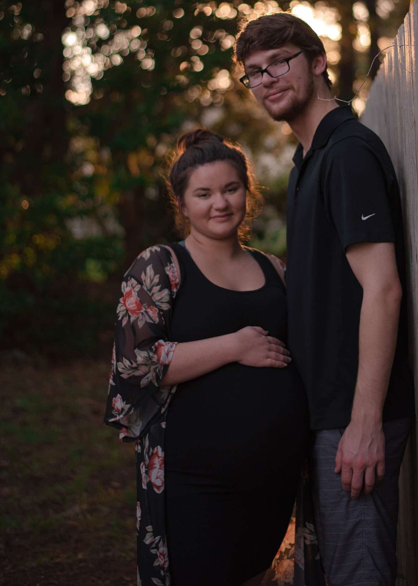 Expectant woman in black dress and floral kimono cradles her baby bump beside a bearded man in glasses at dusk