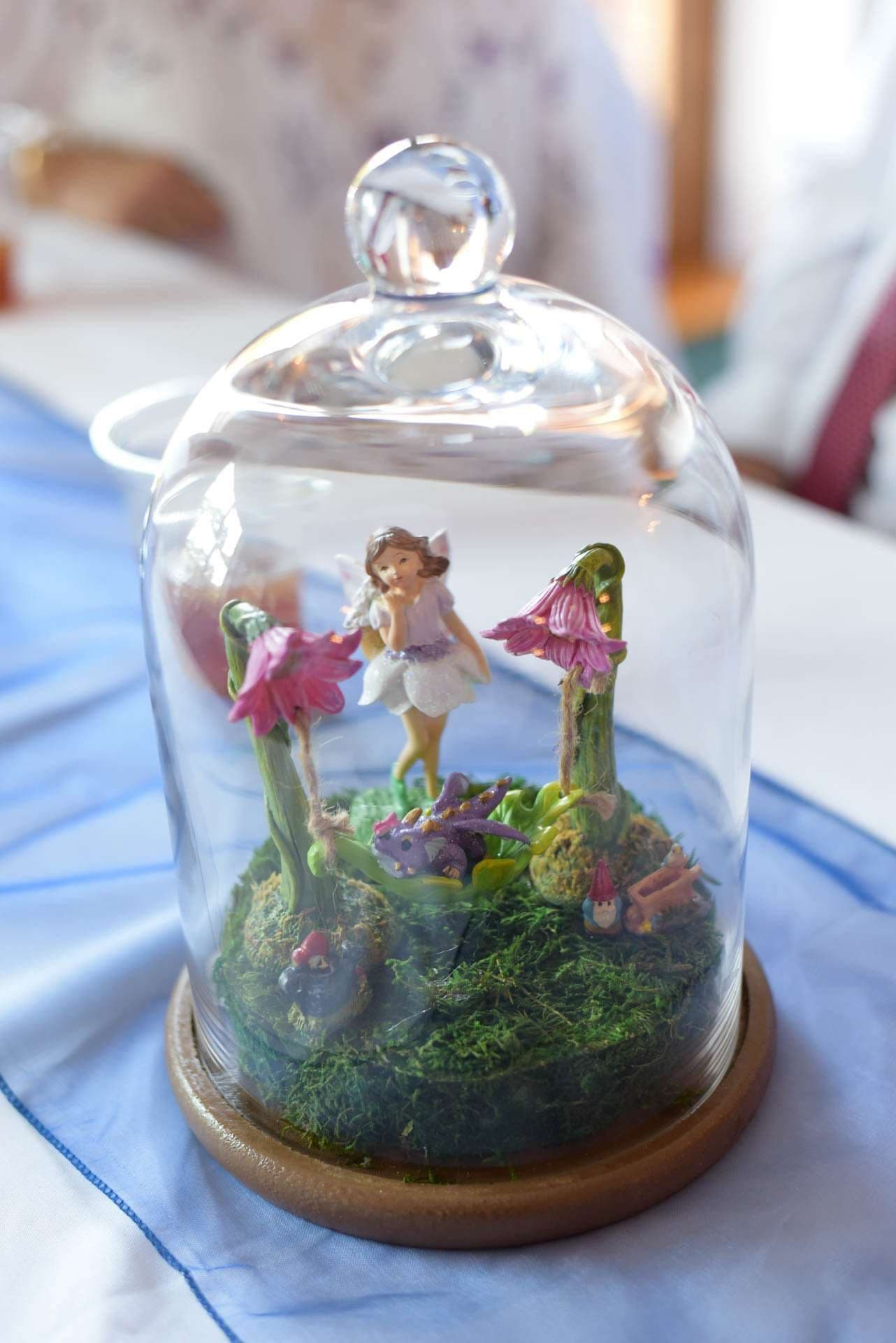 Glass cloche on a wooden base holding a tiny fairy figurine among pink flowers and moss on a blue table runner