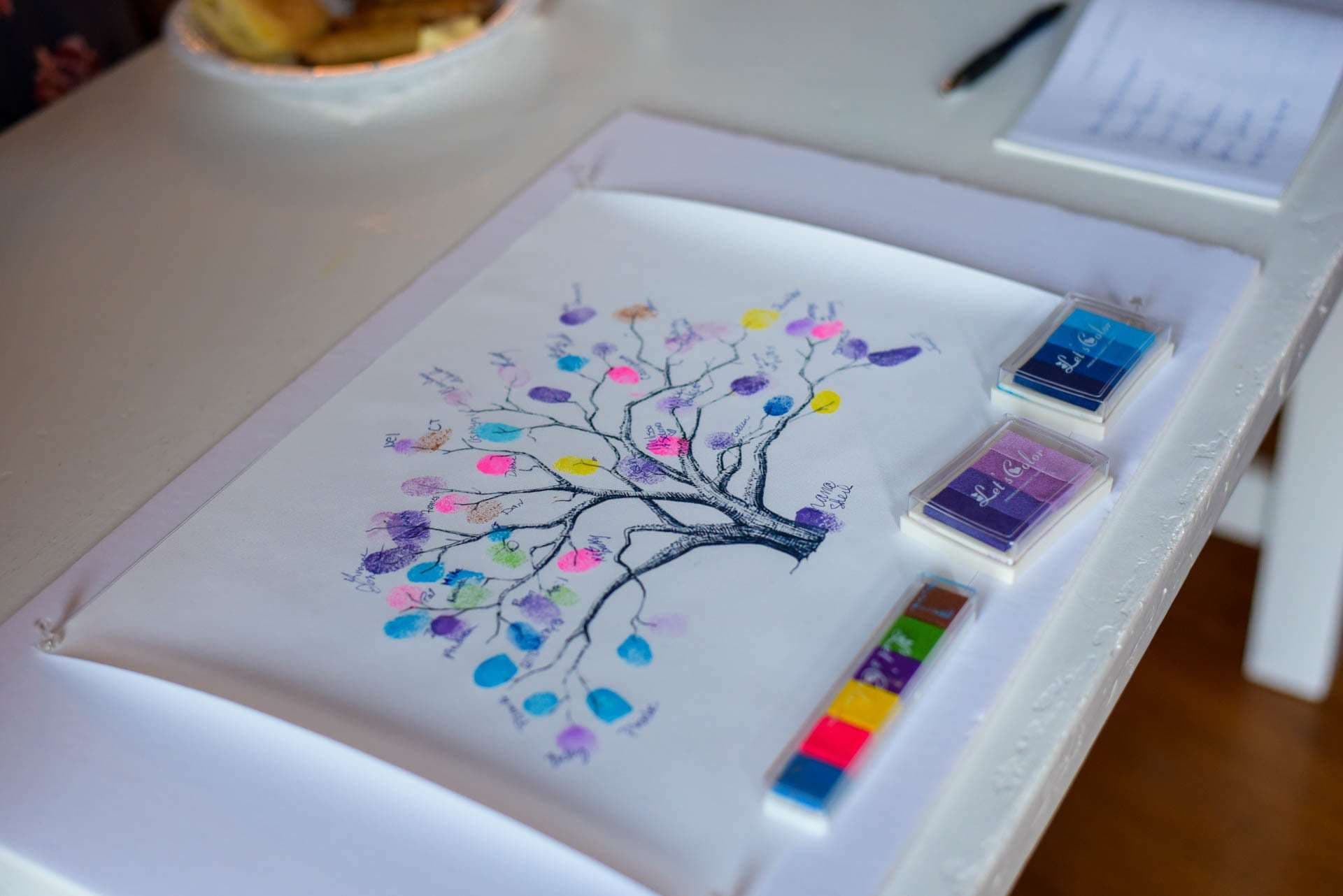 Fingerprint guest book tree with colorful thumbprint leaves on a white table beside blue, purple and rainbow ink pads