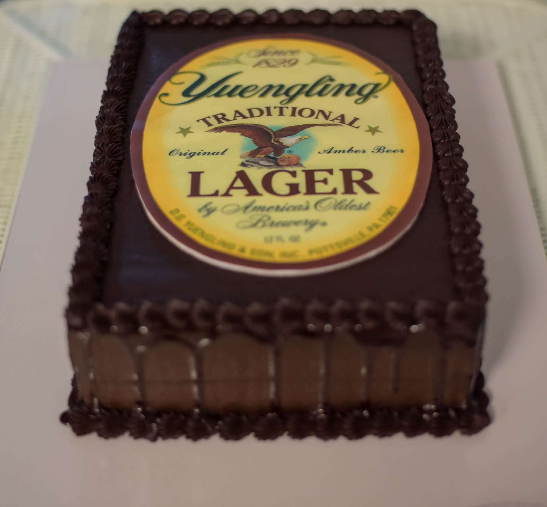 Rectangular chocolate cake with dark piped border topped with a Yuengling Traditional Lager label on a white board