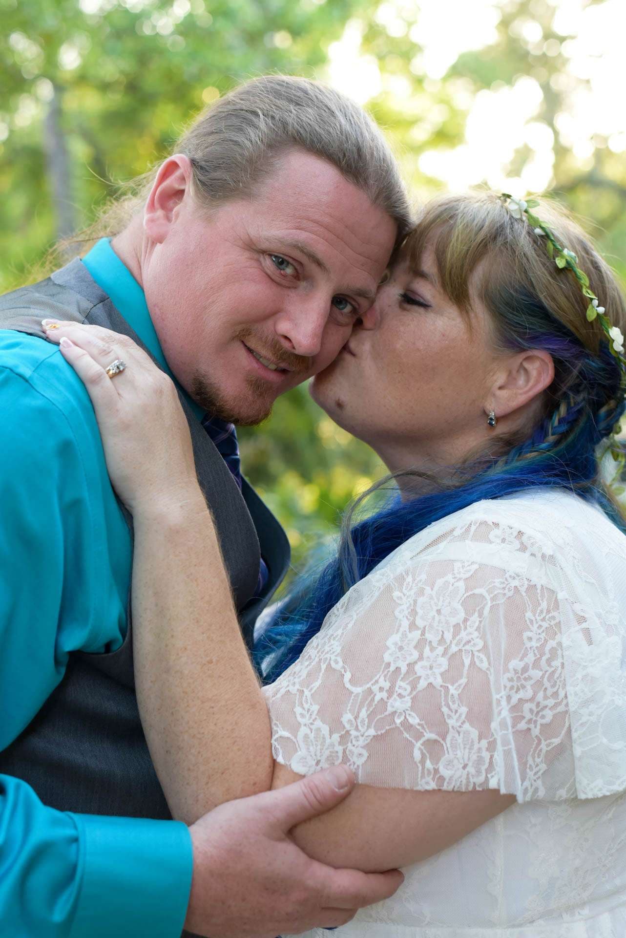 Woman in white lace dress with blue hair and flower crown kisses the cheek of a smiling man in teal shirt