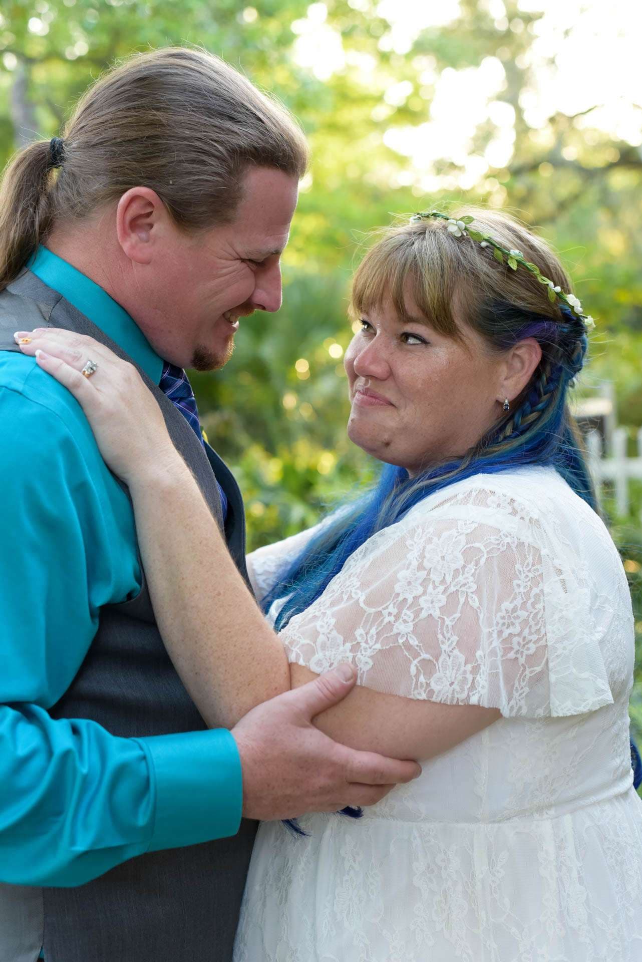 Smiling couple embracing outdoors, the bride in a white lace dress with blue hair and a flower crown