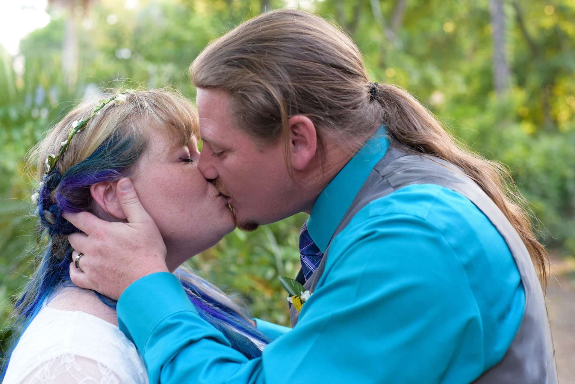 Couple kissing outdoors among greenery, she has blue and purple hair with a flower crown, he wears a teal shirt and gray vest