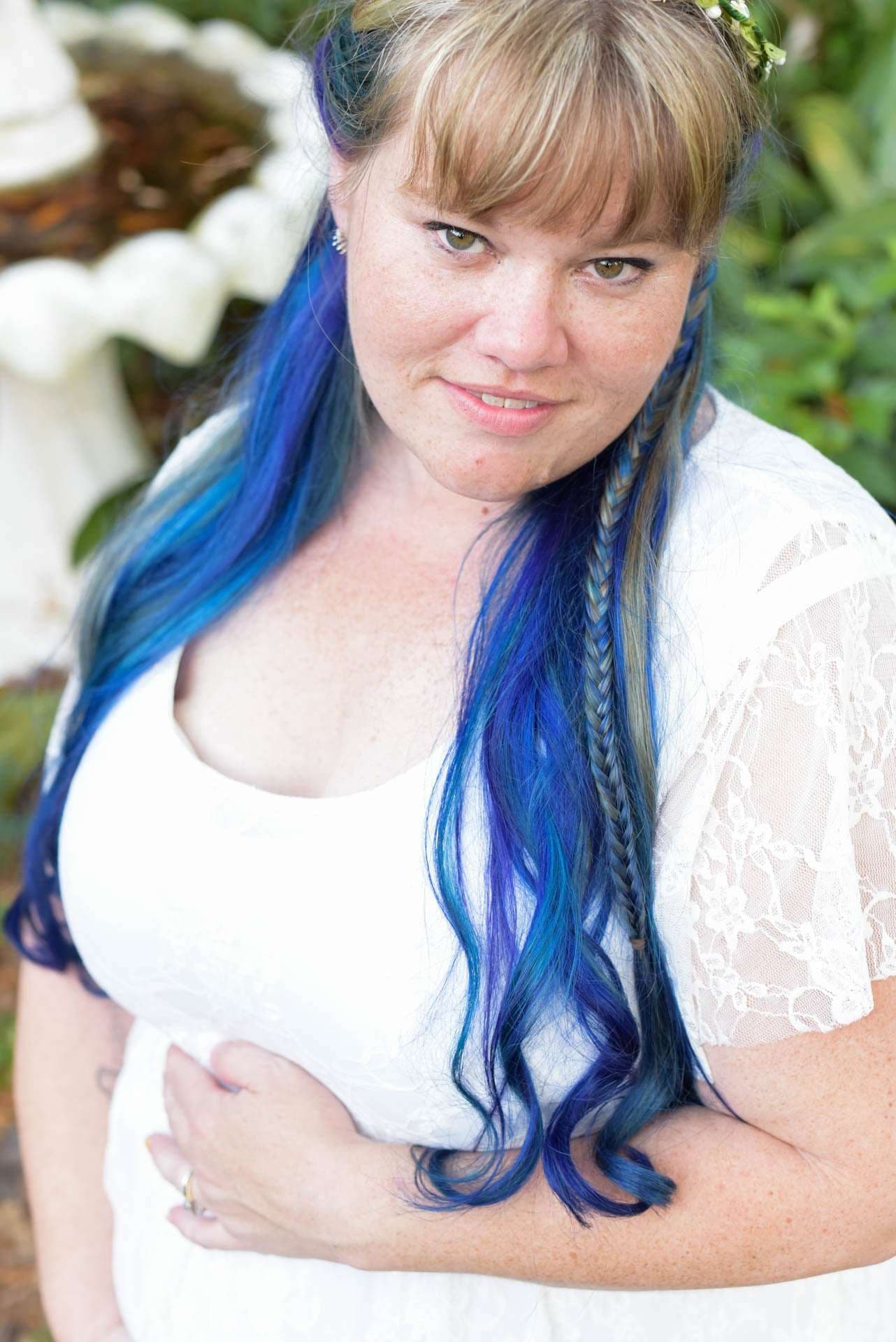 Woman with long blue and purple hair and a side braid, wearing a white lace dress, smiling in a garden