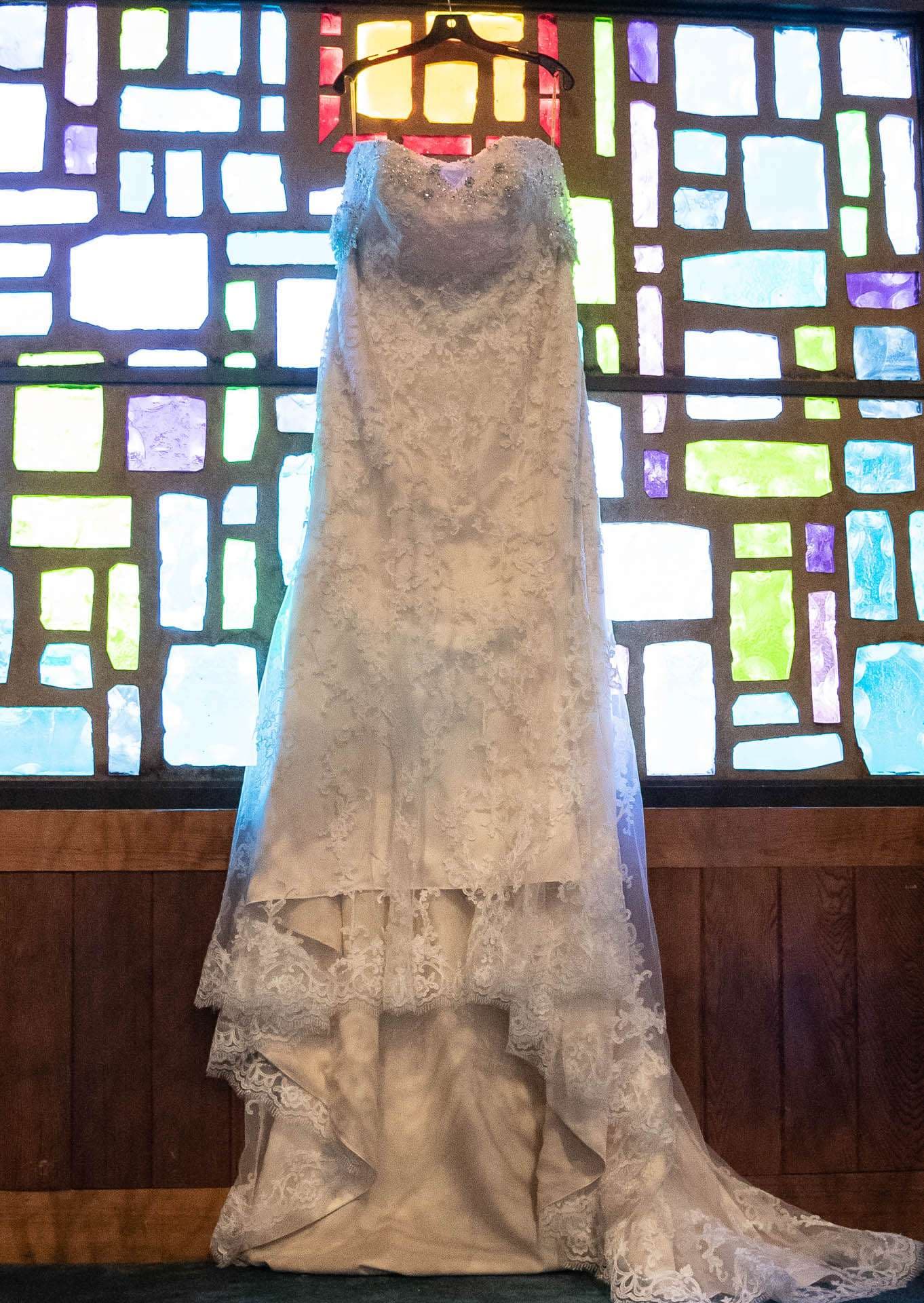 Strapless white lace wedding gown hanging in front of a colorful stained glass window above wood paneling